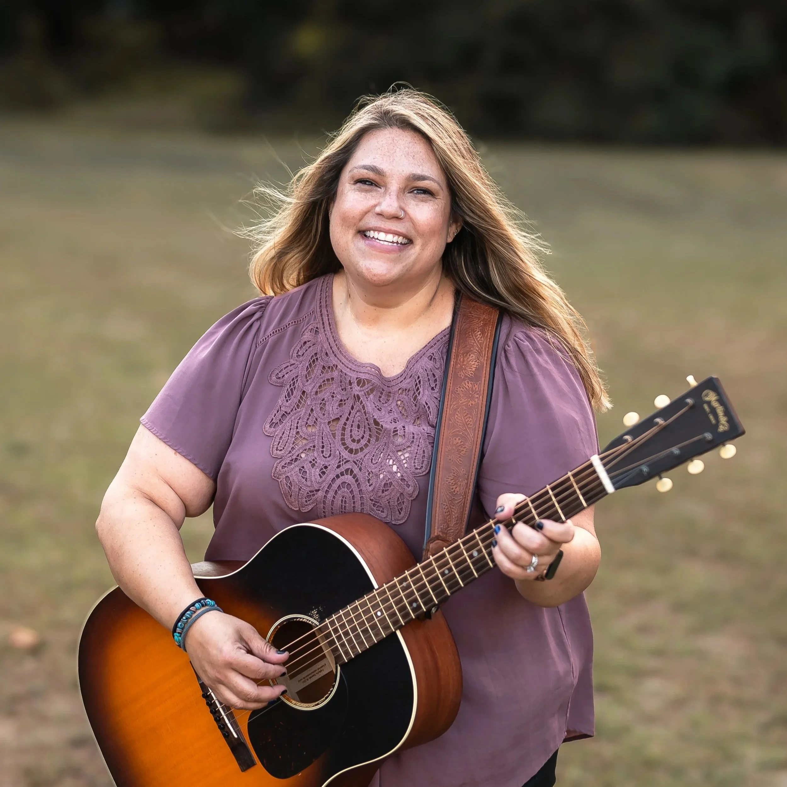 Beka Jones Music with long wavy hair playing acoustic guitar outdoors at sunset near John Wright's Restaurant, wearing an embroidered purple blouse.