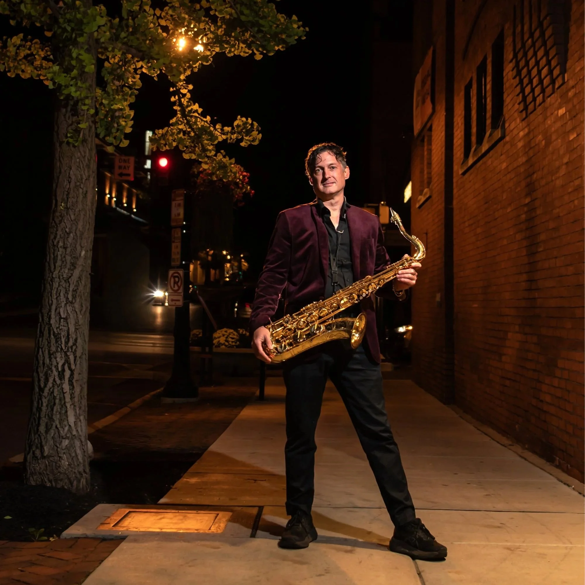 Saxophonist in a purple jacket performing at night outside Southern Market, Lancaster PA, illuminated by streetlight with brick buildings in the background.
