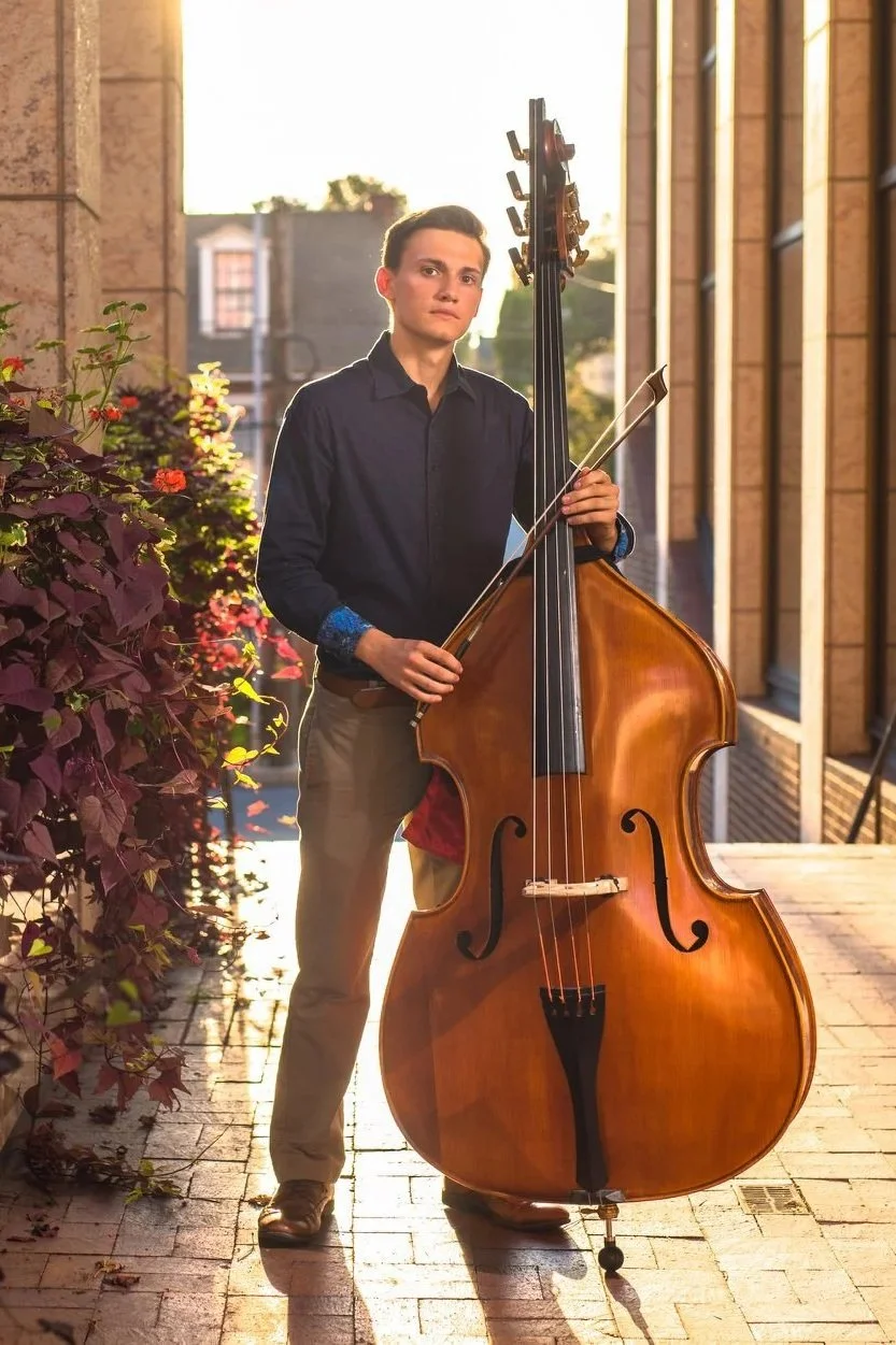 A young man standing outdoors on a brick sidewalk at sunset at the WARE center in Lancaster PA, holding a double bass and a bow, with building windows and a wall with plants beside him.