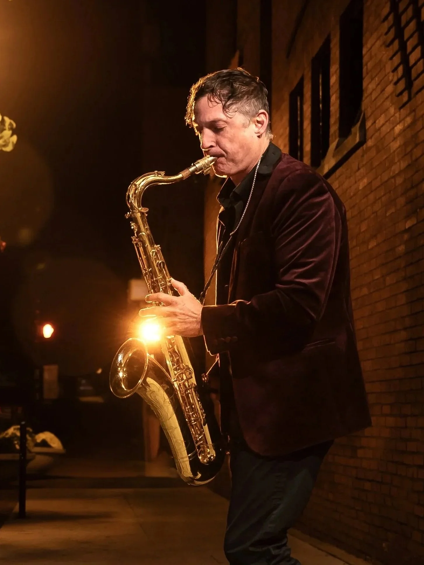 A man is playing a saxophone on a dimly lit street at night, with warm lighting from a nearby streetlamp and a brick wall in the background.