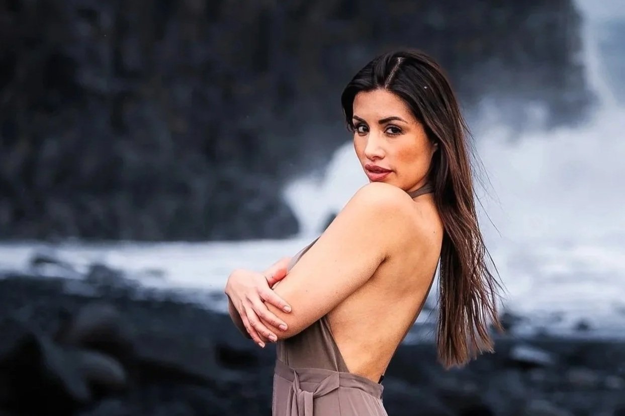 Michelle Benitez, Univision news anchor, stands on black volcanic rocks on an Iceland beach, dark cliffs and crashing waves behind her, looking back over her shoulder in a backless dress.