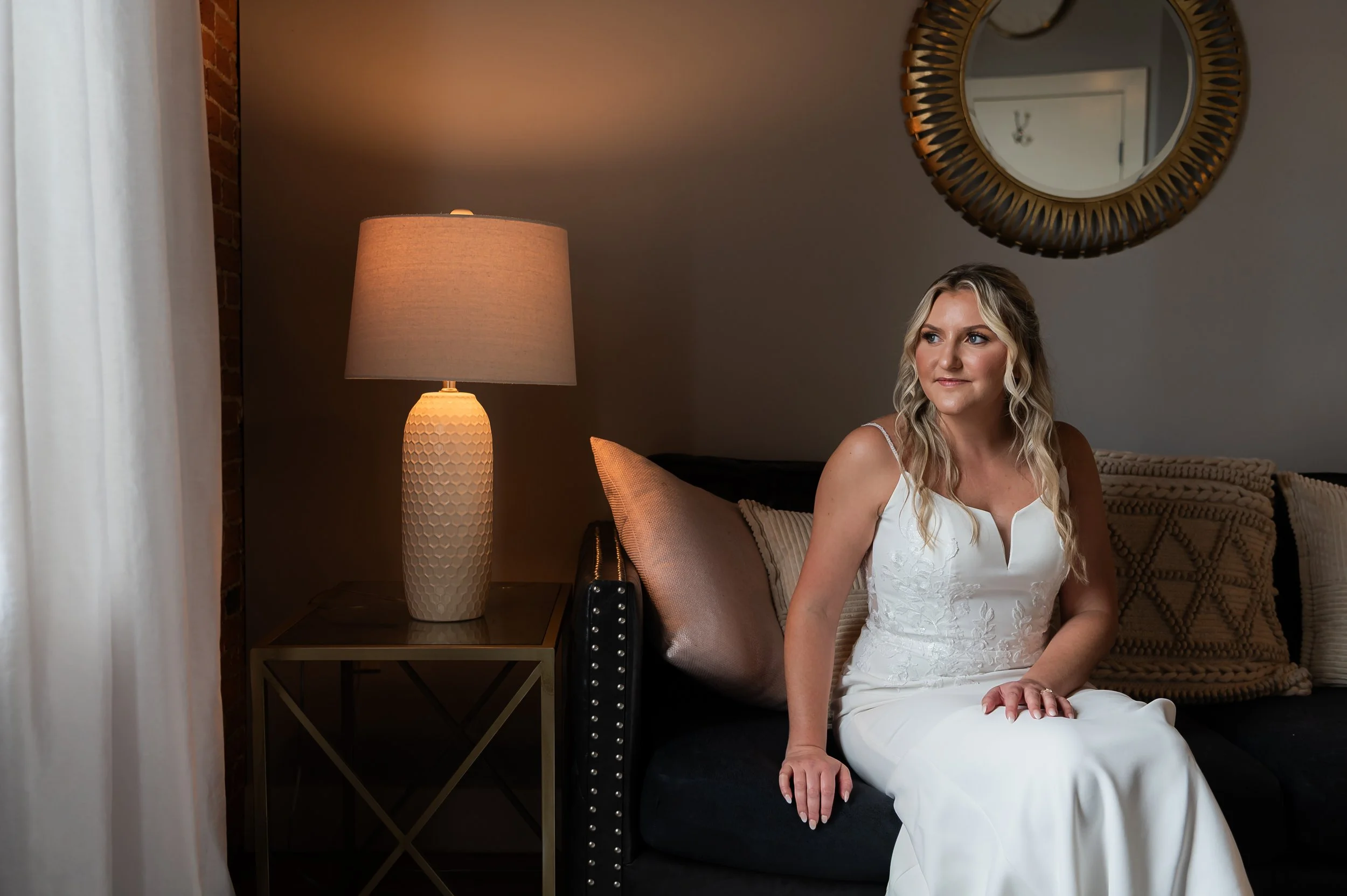 Bride in a white dress sitting on a velvet sofa at The Booking House, Manheim PA, brick wall and decorative mirror nearby.