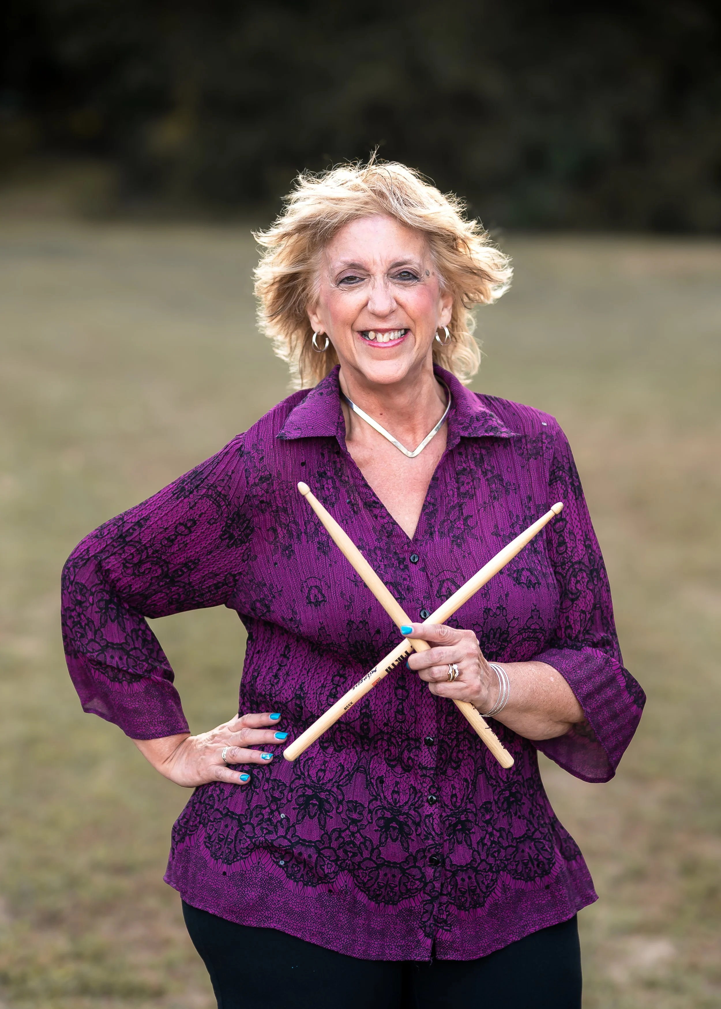 Smiling blonde woman from Winged Victory Band holding drumsticks crossed in front of her chest outdoors near John Wright's Restaurant, with a blurred grassy background.