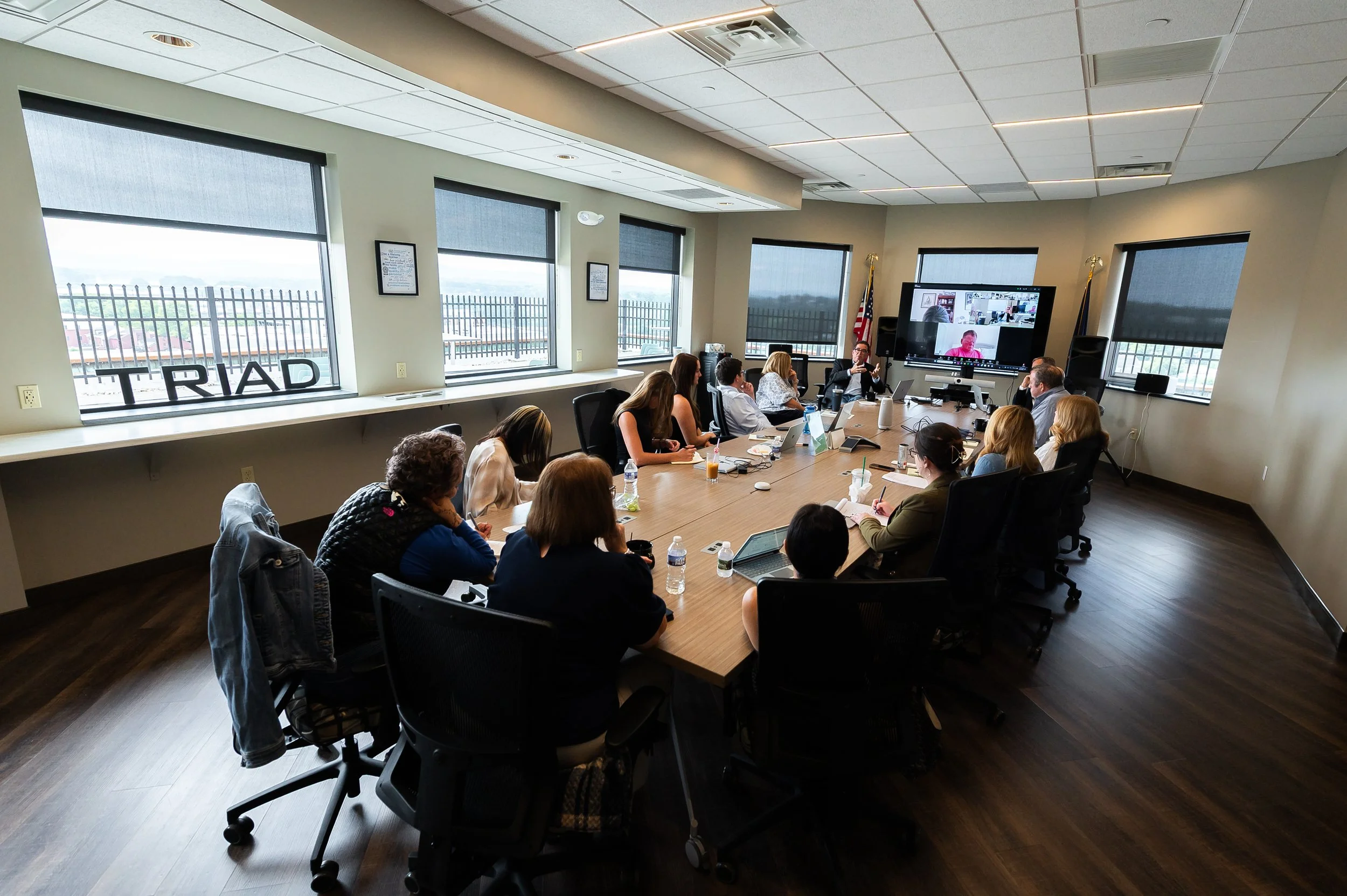 A group of people attending a meeting in a conference room with large windows, some working on laptops, and a video conference call displayed on a large screen at the front of the room.