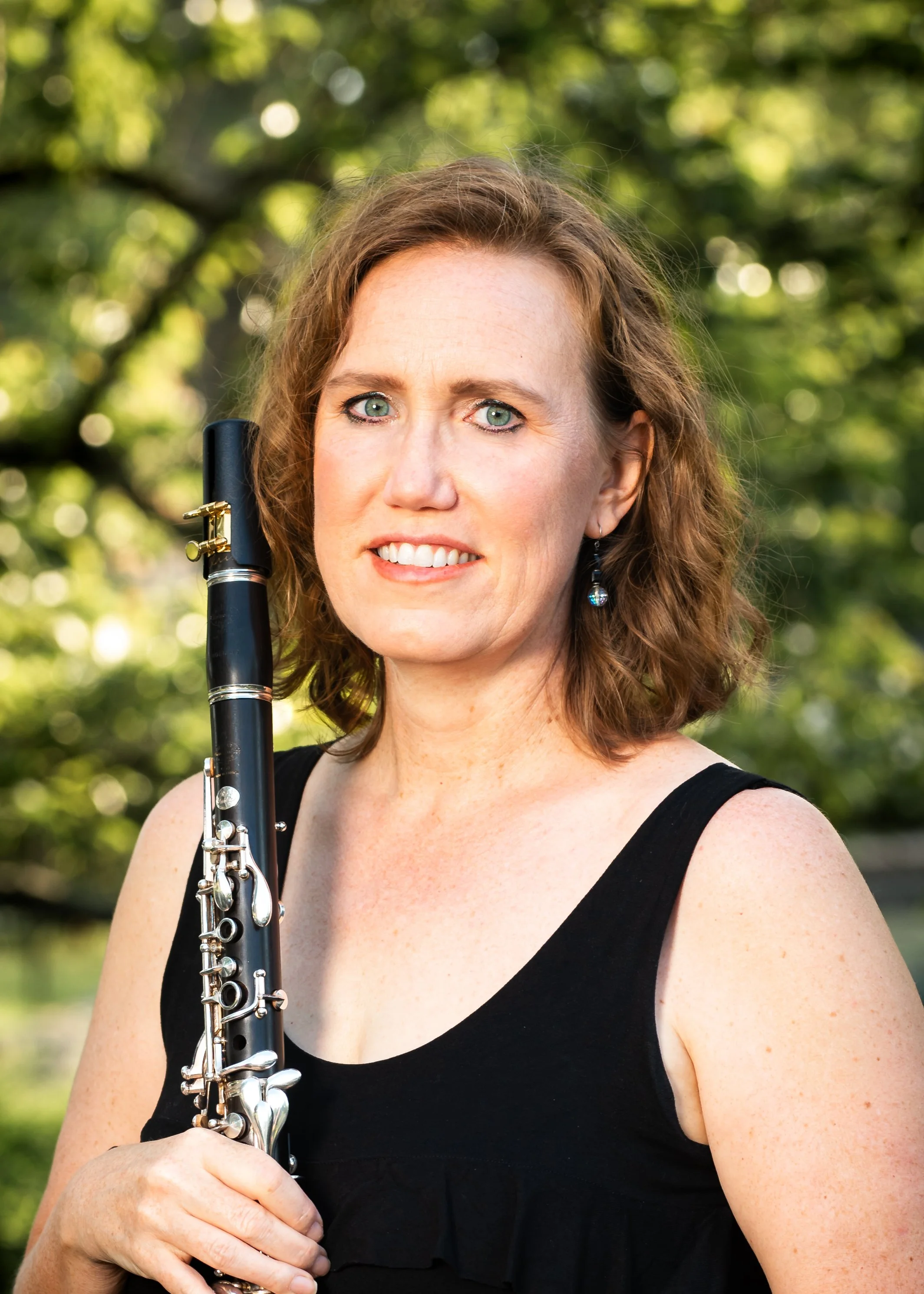 A woman with curly brown hair and blue eyes holding a black flute outdoors with a background of green trees and sunlight.