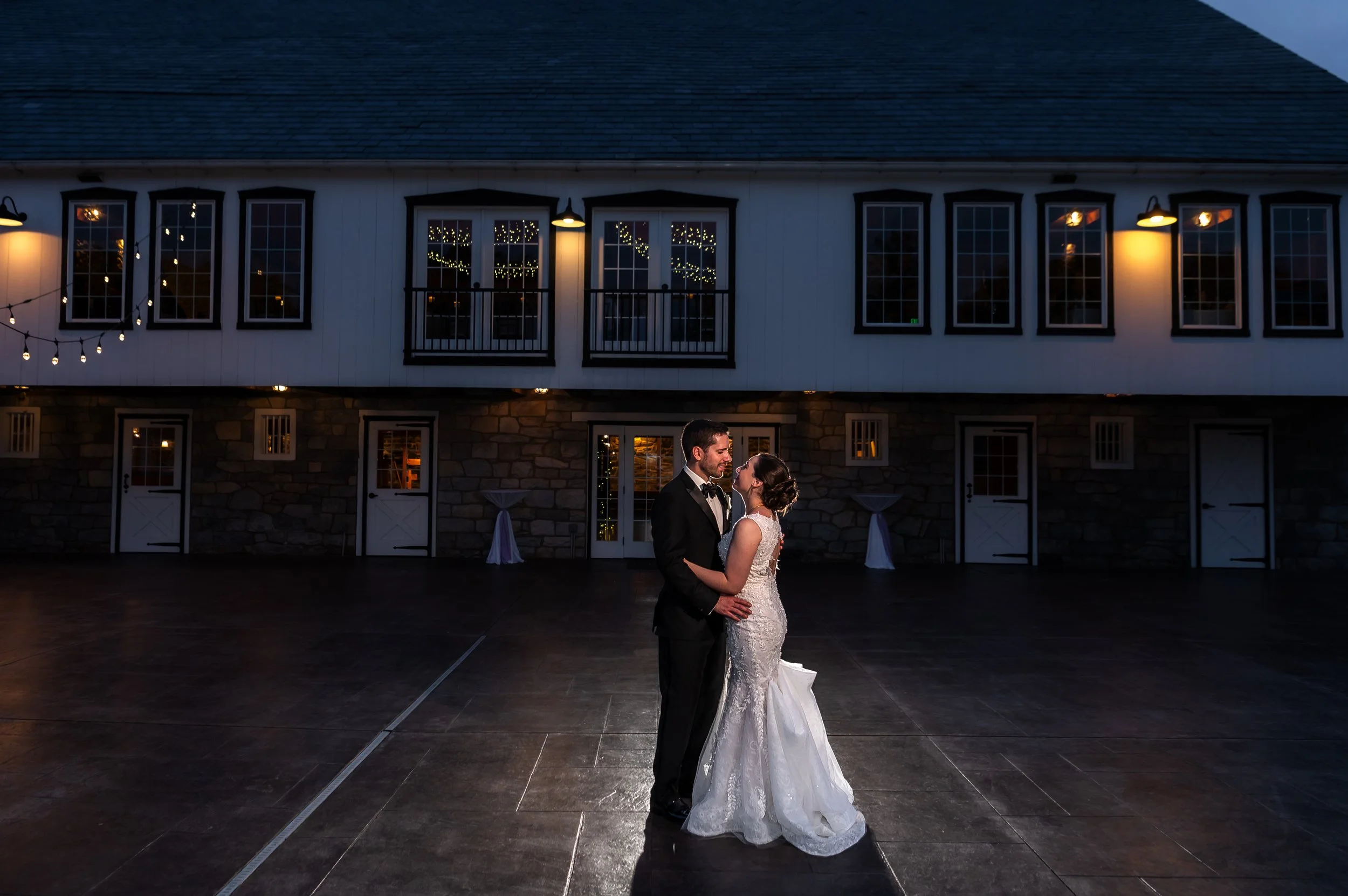 Bride and groom dancing closely at The Barn at Silverstone, Lancaster PA, warm string lights on the venue's exterior wall behind them.