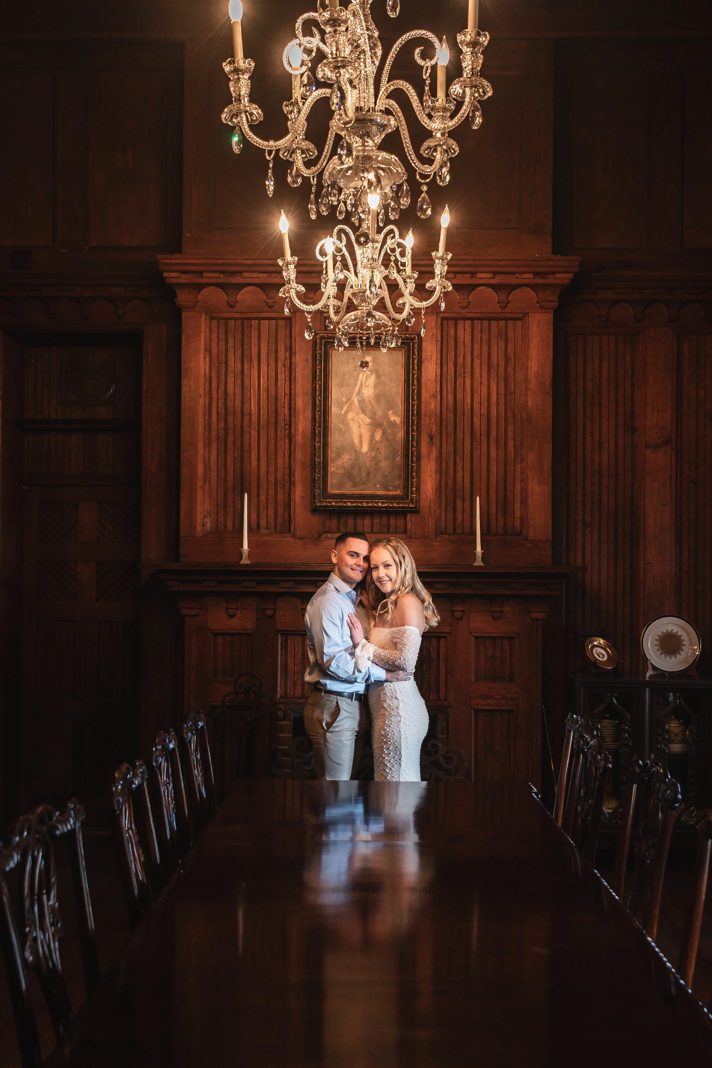 Couple embracing in a wood-paneled room at Berkeley Castle, West Virginia, ornate chandelier overhead, bride in an off-the-shoulder white dress.