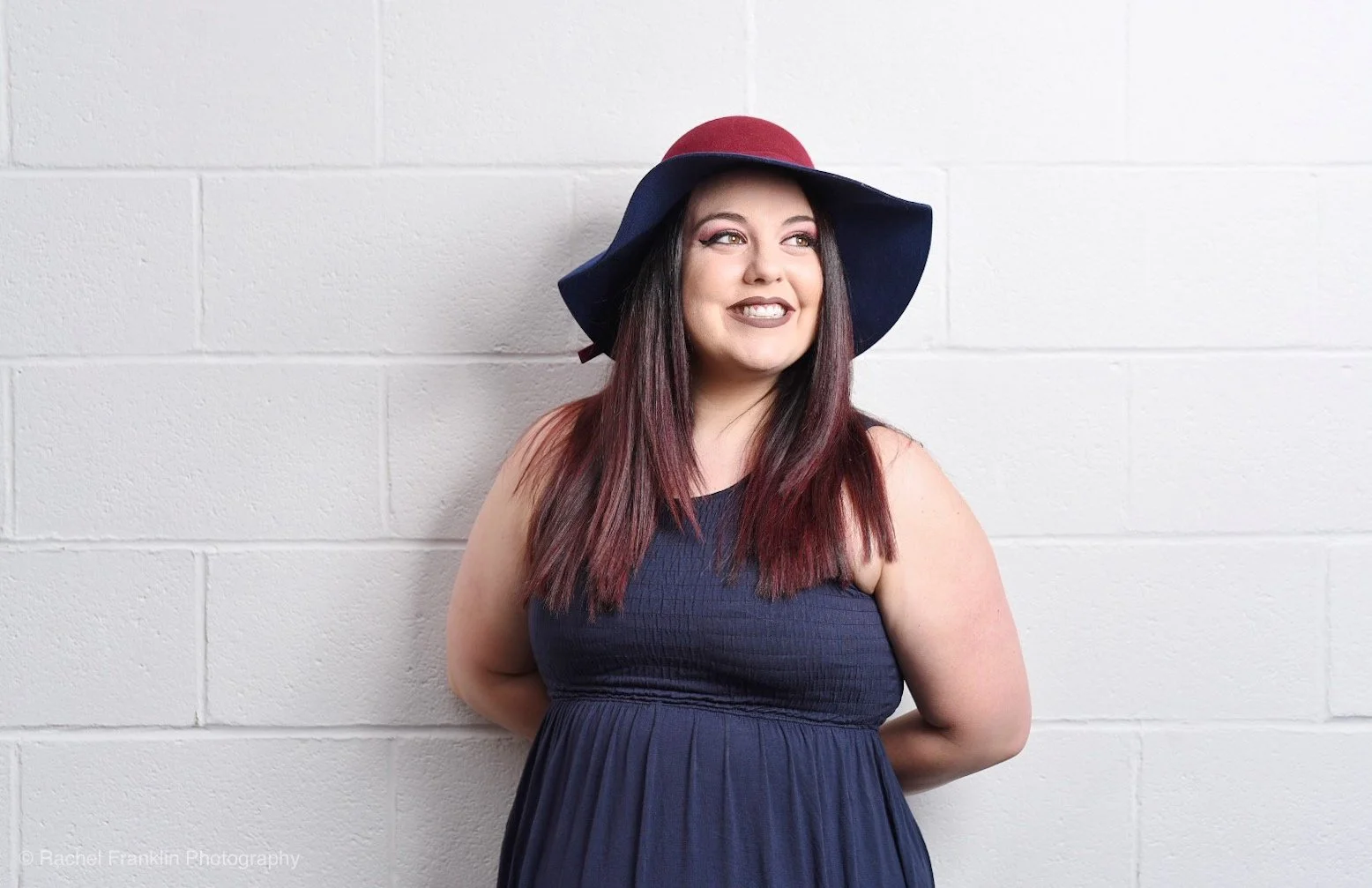 A woman with long brown hair wearing a large navy blue and red floppy hat and a navy blue sleeveless dress, standing against a white brick wall, smiling and looking slightly to the side.