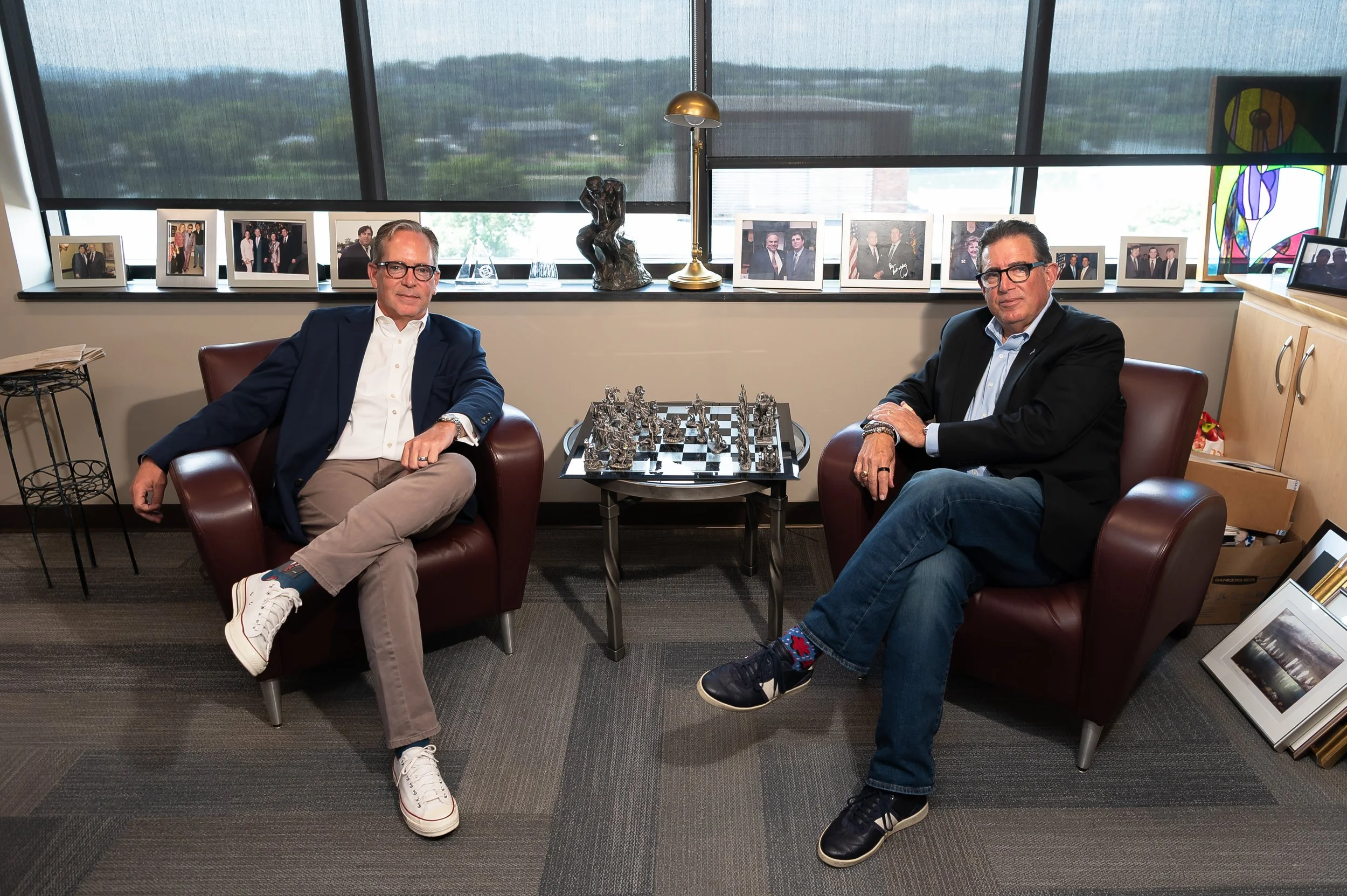Two men sitting in leather armchairs in an office with a windowed background, decorated with framed photographs, sculptures, a brass lamp, and colorful artwork.