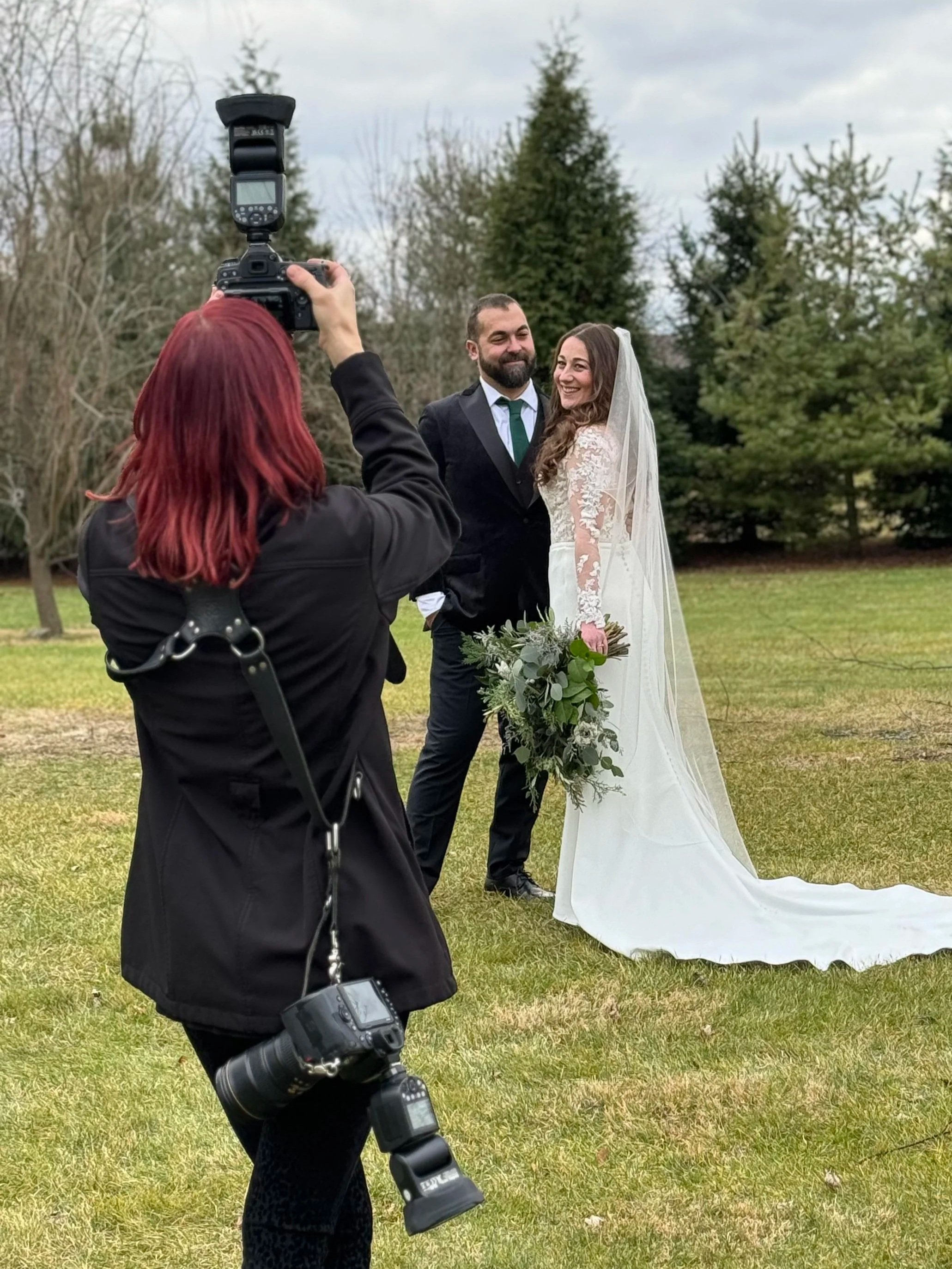 A bride and groom posing outdoors at Farm 1896 on their wedding day, with the bride holding a bouquet of white flowers and greenery, surrounded by wedding guests and a house with a red roof in the background.