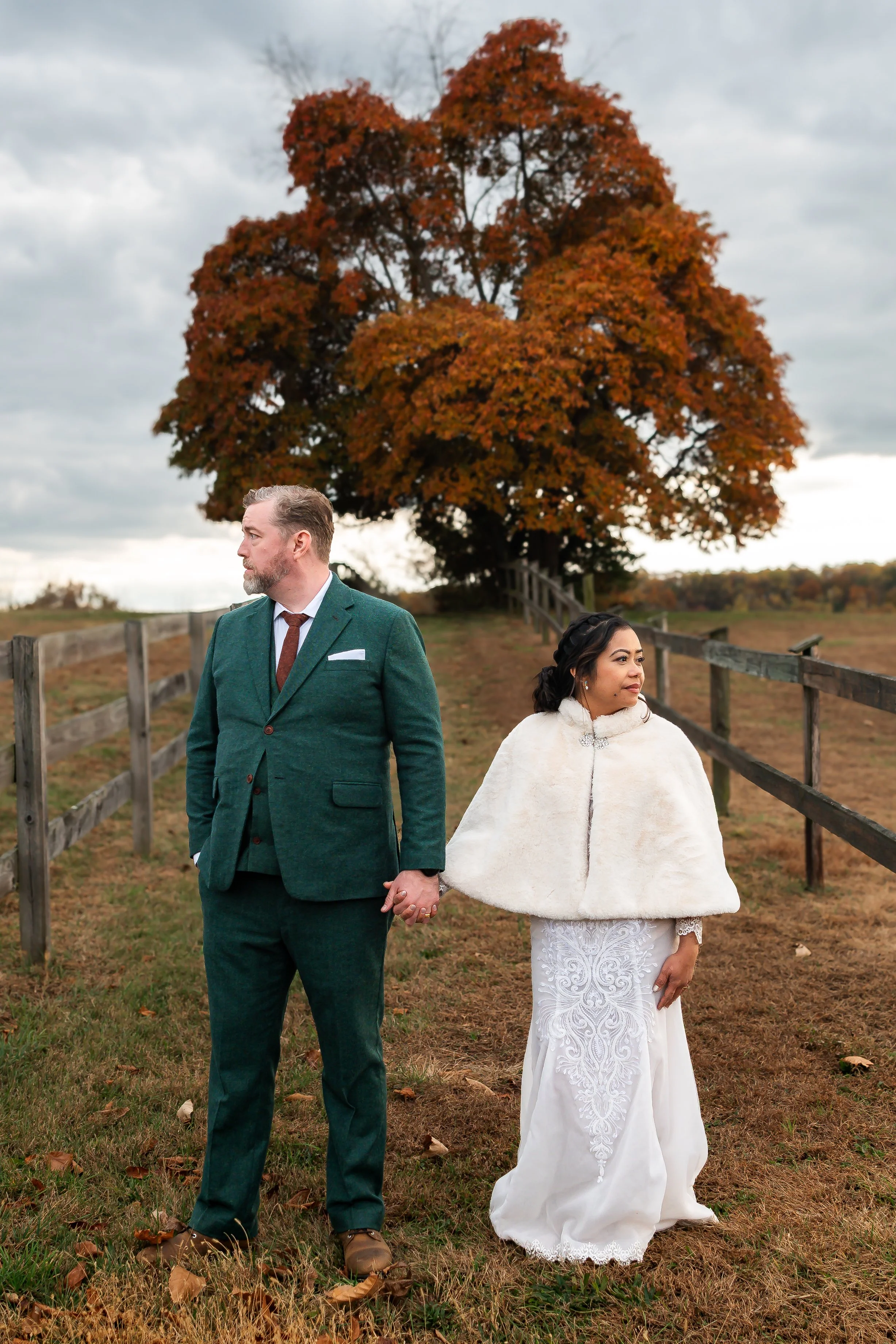 A bride and groom hold hands while standing outdoors on a grassy area with fallen leaves, near a wooden fence, under a large tree with orange and red autumn leaves, with overcast skies in the background.