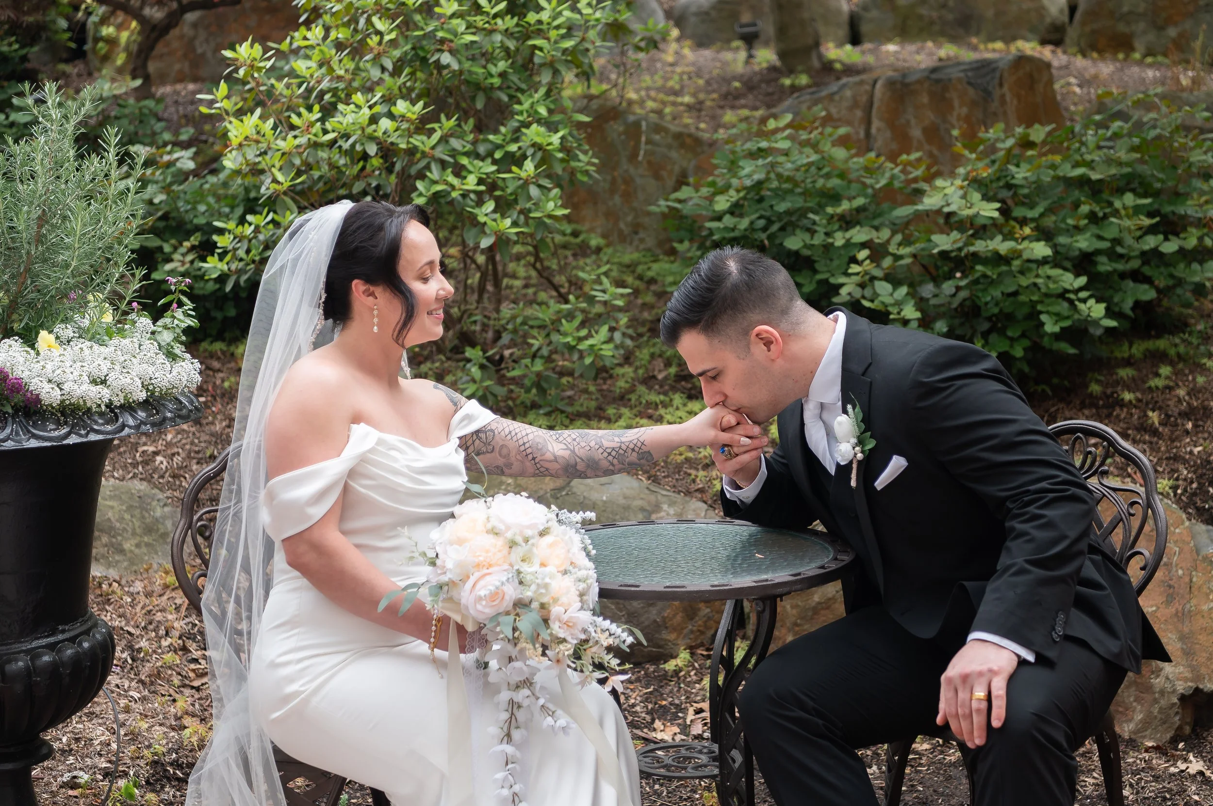 Groom kissing bride's hand at an outdoor table at Stone Gables Estate's Star Barn, Elizabethtown PA, surrounded by greenery and potted flowers.