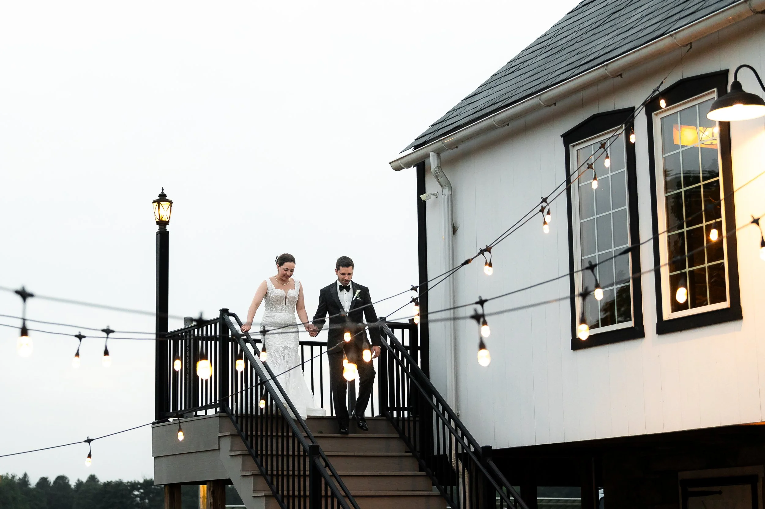 Bride and groom holding hands on a balcony at dusk at The Barn at Silverstone, Lancaster PA, string lights and a lamppost nearby.