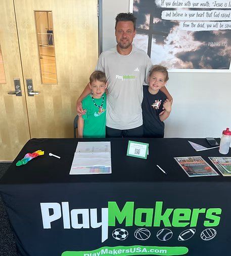 Man standing with two young girls behind a PlayMakers table at an indoor event.