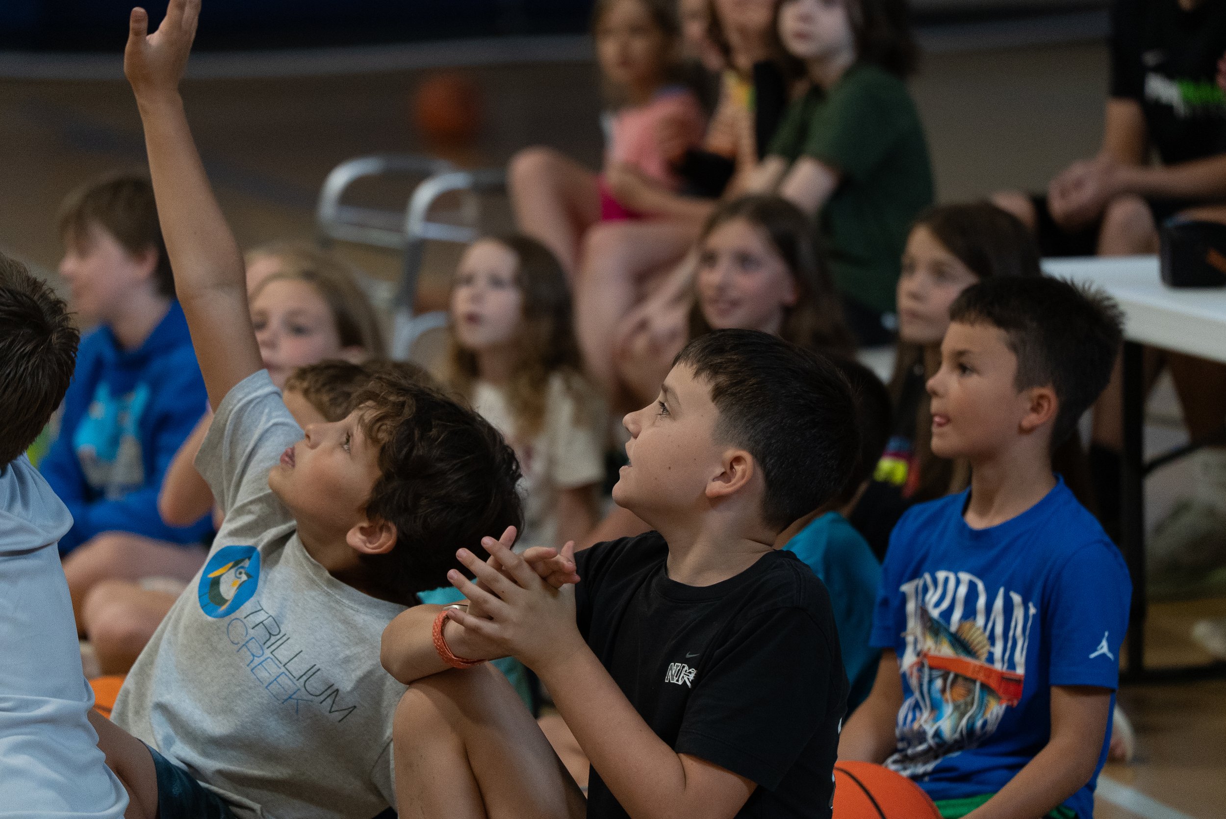 Group of children sitting and engaging during an indoor event, with one child raising his hand and others smiling and interacting.