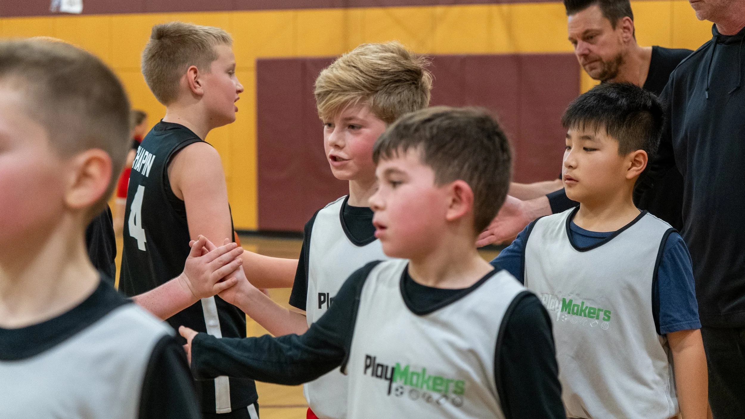 Young boys in sports jerseys shaking hands after a game in an indoor gym, with two adult men overseeing.