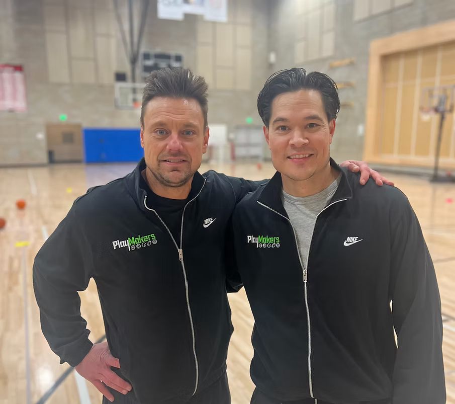 Two men in black Nike jackets with 'PlayMakers' logos on the chest, standing side by side in a gymnasium, smiling, with one arm around each other's shoulders.