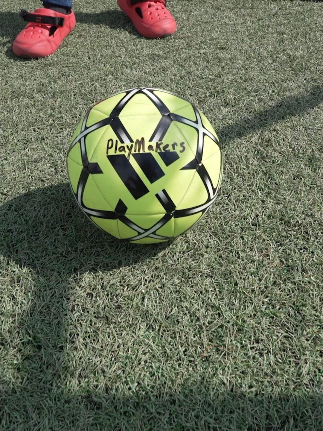 A yellow and black soccer ball with the words 'Play Makers' written on it, placed on green grass. There are red shoes of a person standing nearby.