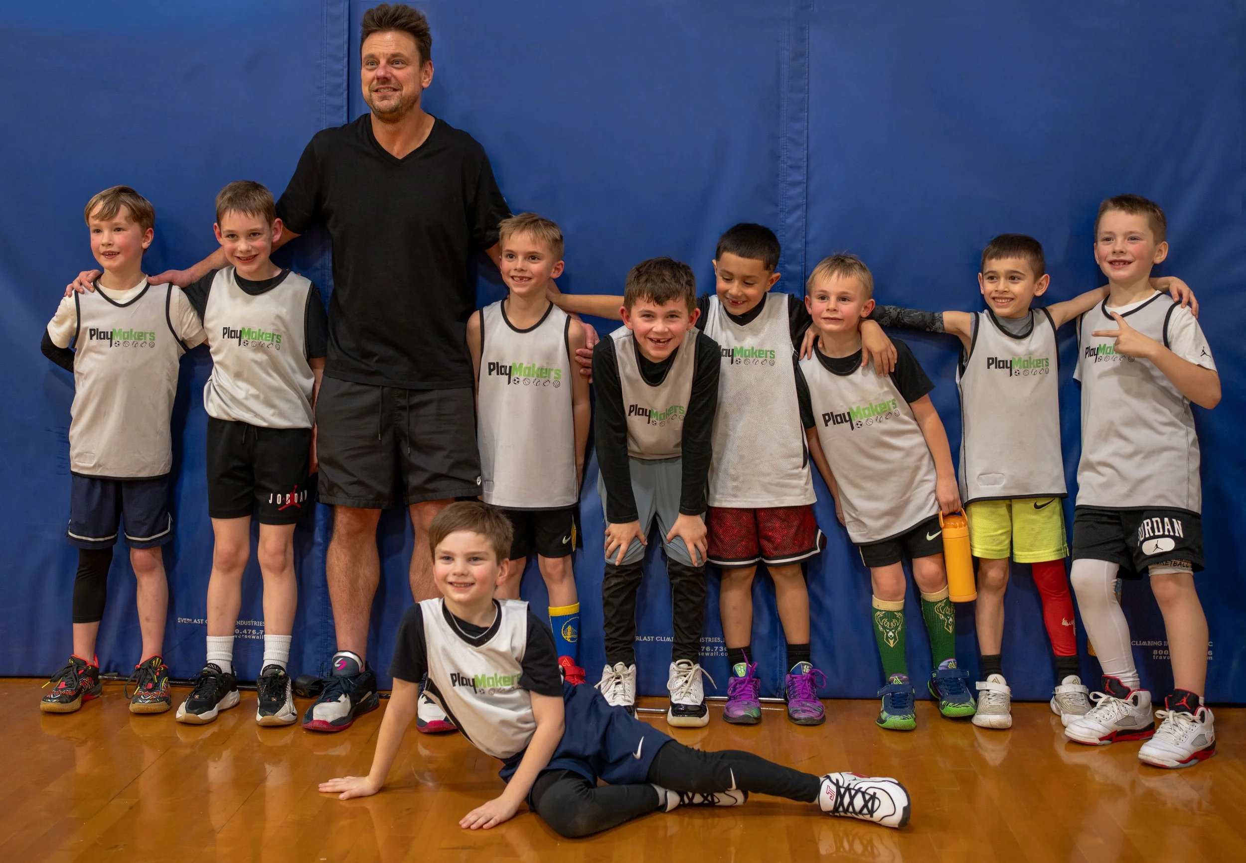 A group of young boys and one adult man posing for a photo in a gymnasium. The boys are wearing sports jerseys with 'PlayMakers' printed on them, and the man is standing behind them with his hands on their shoulders. They are all smiling, and some boys are pointing or have their arms around each other. The gym has a blue padded wall and a wooden floor.
