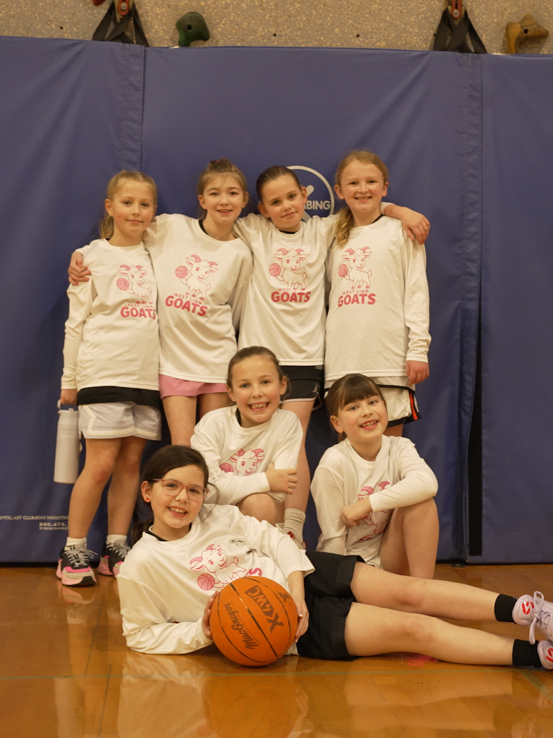 A group of young girls on a basketball team called the West Linn Goats, posing in a gym, wearing matching white shirts with a goat logo, some holding a basketball, with a blue padded wall in the background.