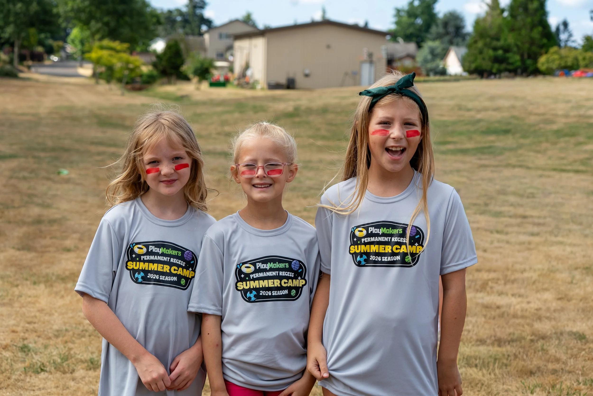 Three young girls with face paint wearing gray T-shirts with summer camp logos, standing outdoors on a grassy area with trees and buildings in the background.