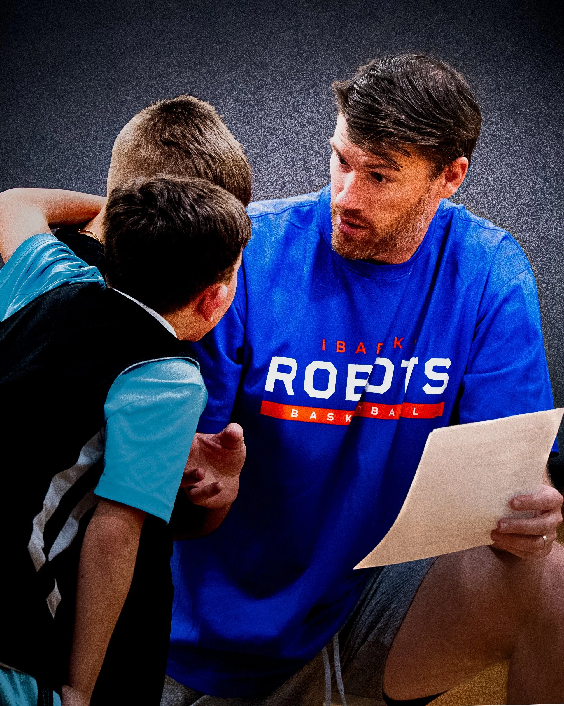 A man in a blue sports jersey with the words 'ROPE'S BASKET' visible, showing a surprised or intense expression, holding a piece of paper, while two children, one with short brown hair and the other with short reddish-blonde hair, are closely engaged in conversation with him inside a room.