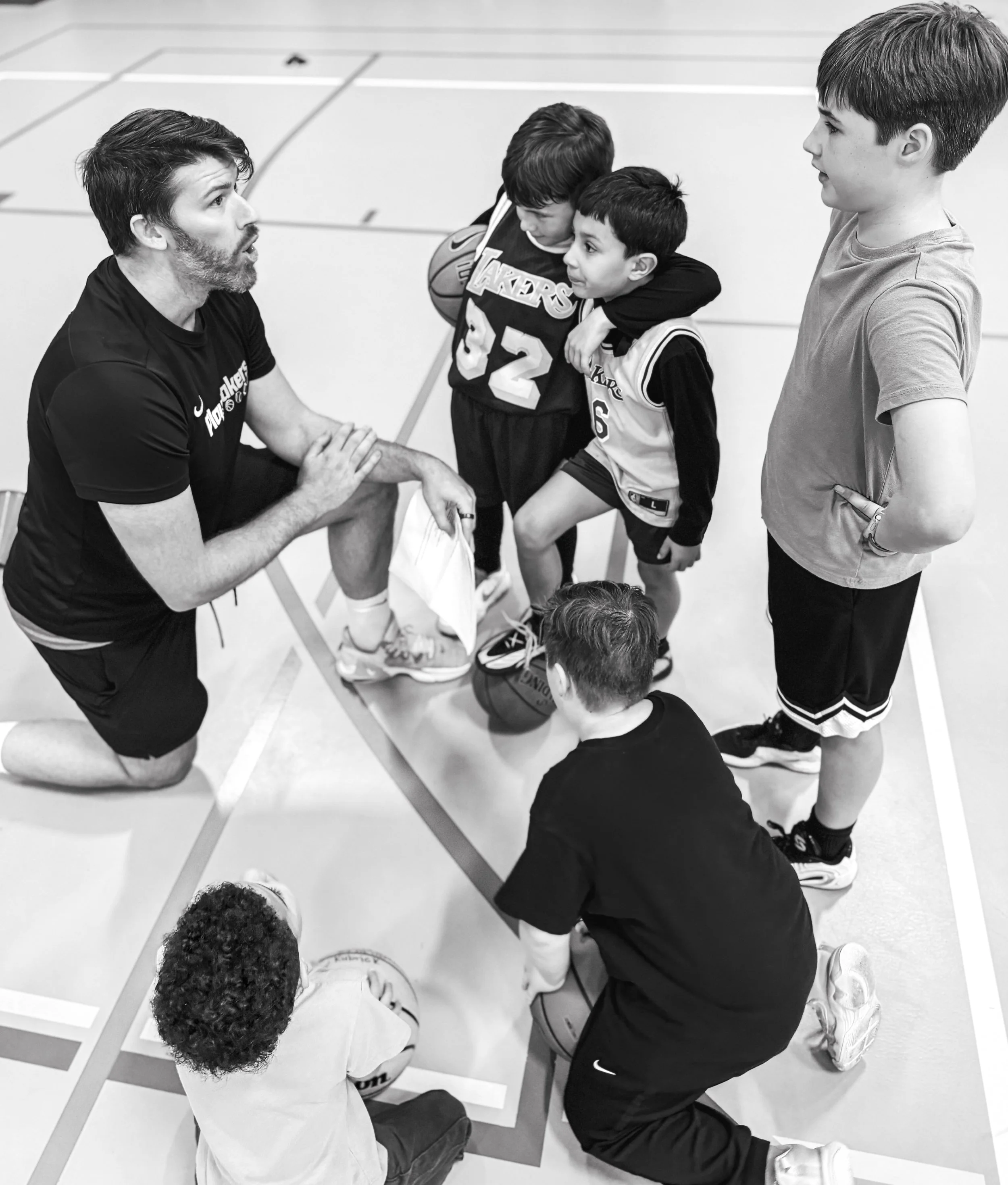 A coach kneeling on a gym floor speaking to a group of young boys who are sitting on the rim of basketballs, wearing sportswear and holding basketballs, in a gymnasium.