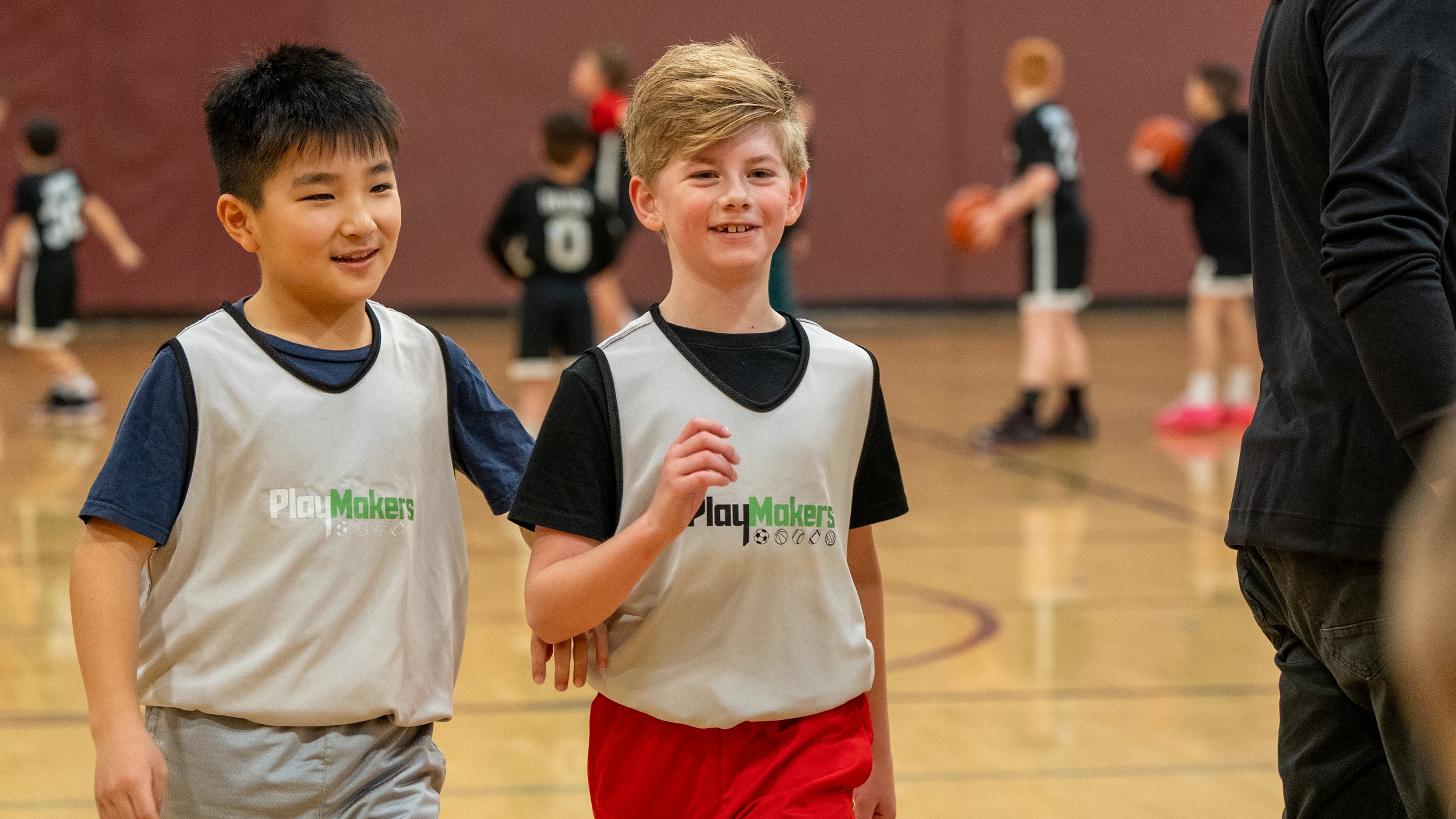 Two boys wearing soccer practice jerseys walking on a basketball court with children playing in the background.