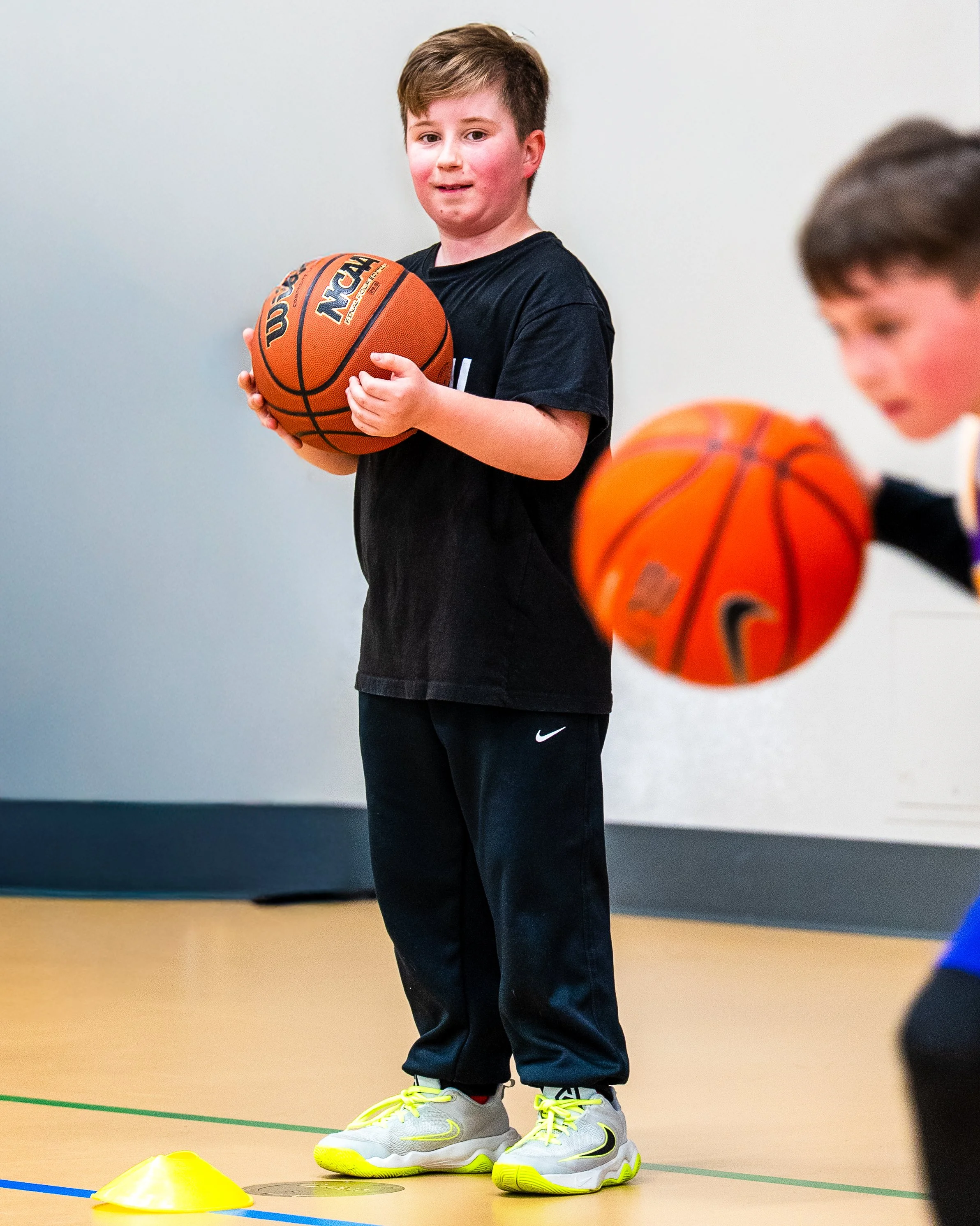 A young boy holding a basketball indoors, near orange cones and a piece of athletic equipment, wearing a black T-shirt, black pants, and yellow and gray sneakers.