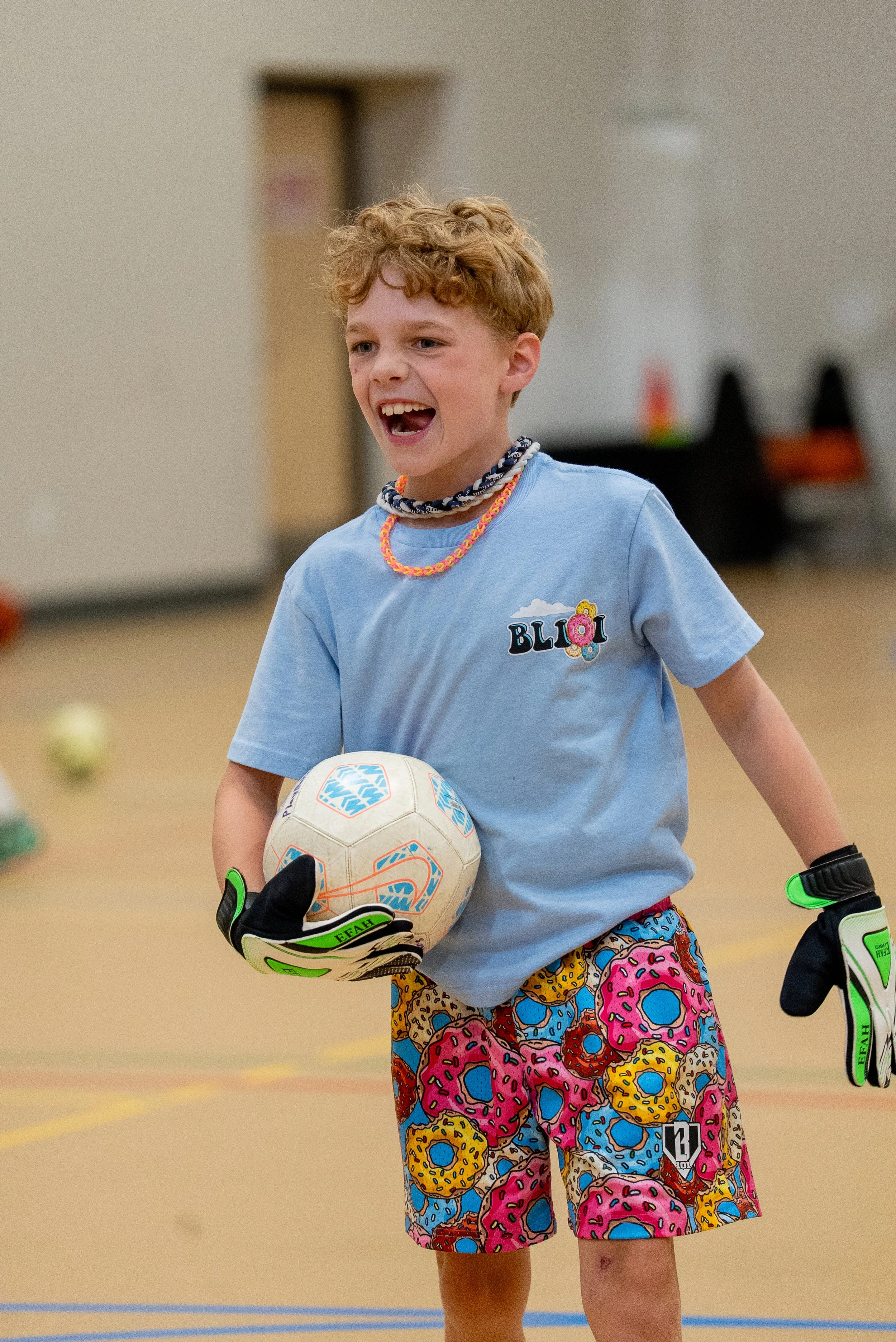A young boy with curly red hair playing indoor soccer, wearing colorful donut-themed shorts, a light blue T-shirt with 'BLOI' on it, and soccer gloves, holding a soccer ball.