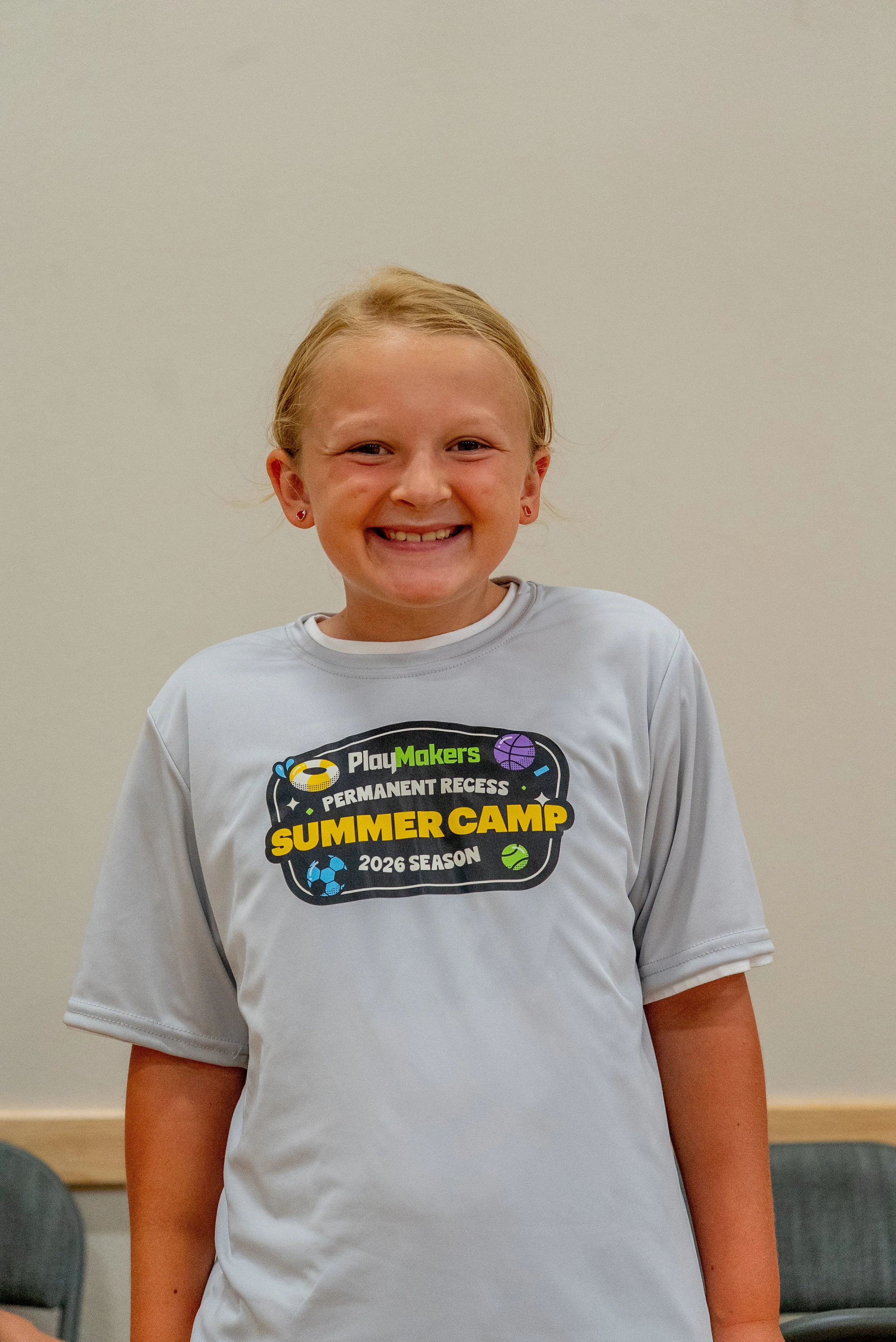 A young girl with blonde hair smiling, wearing a white T-shirt for a summer camp.