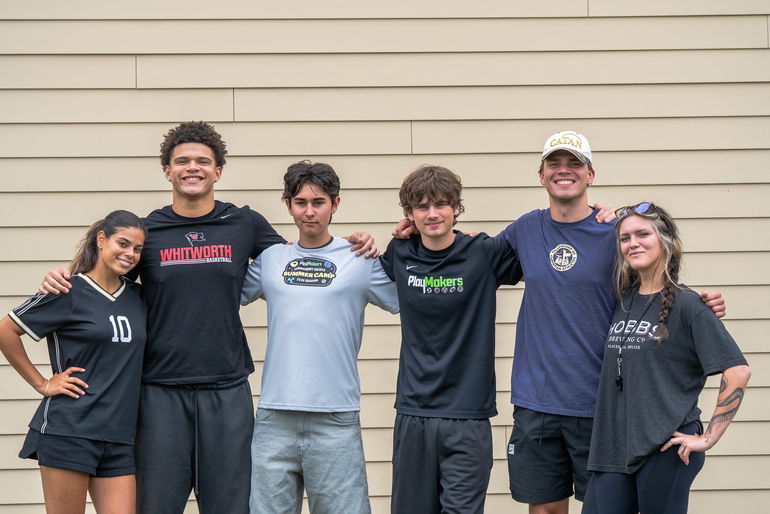 Group of six young people standing with arms around each other against a beige horizontal siding wall, smiling at the camera. Four males and two females, casual athletic and streetwear attire.
