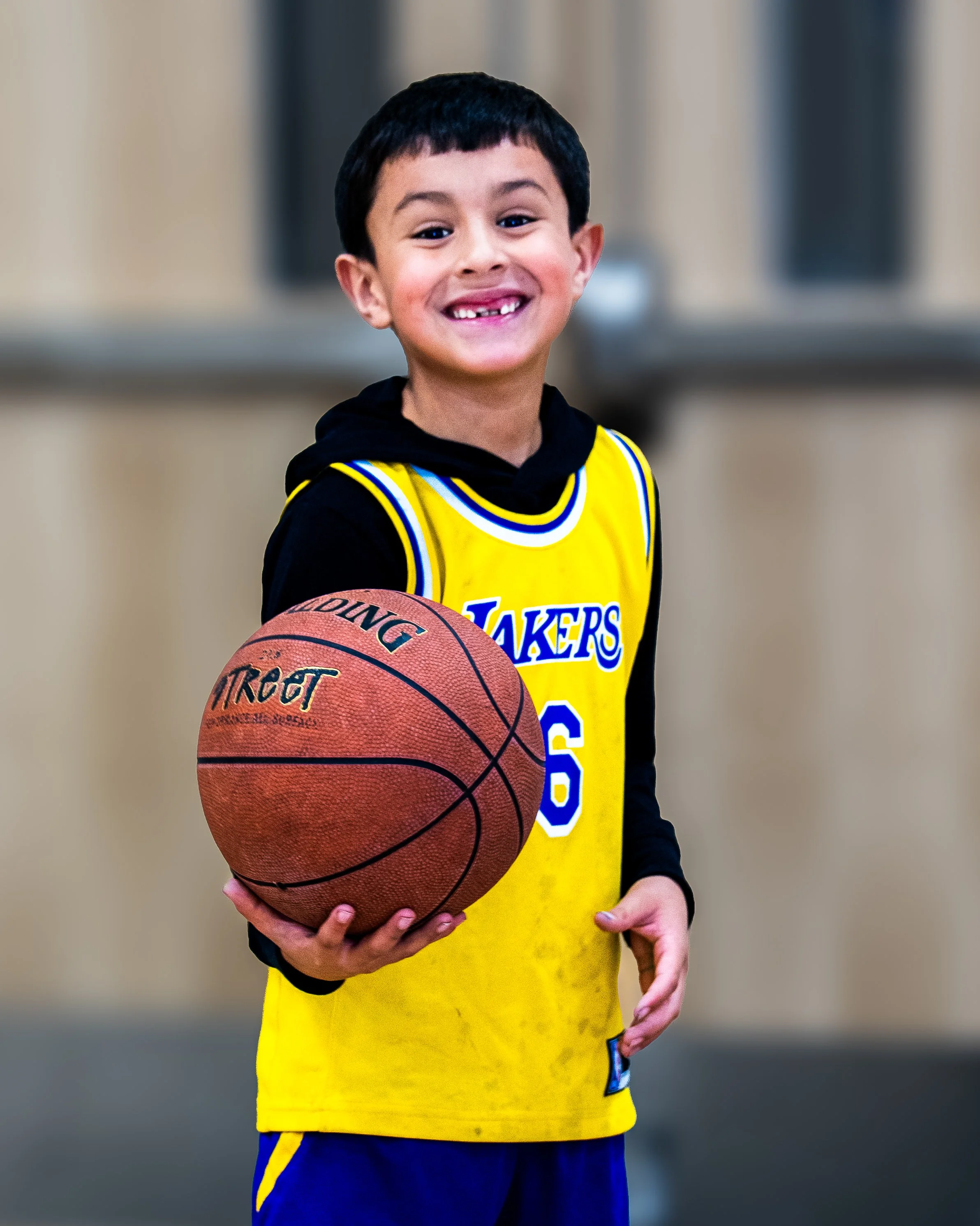 A young boy in a Los Angeles Lakers basketball jersey holding a basketball, smiling, indoors with a blurred background.