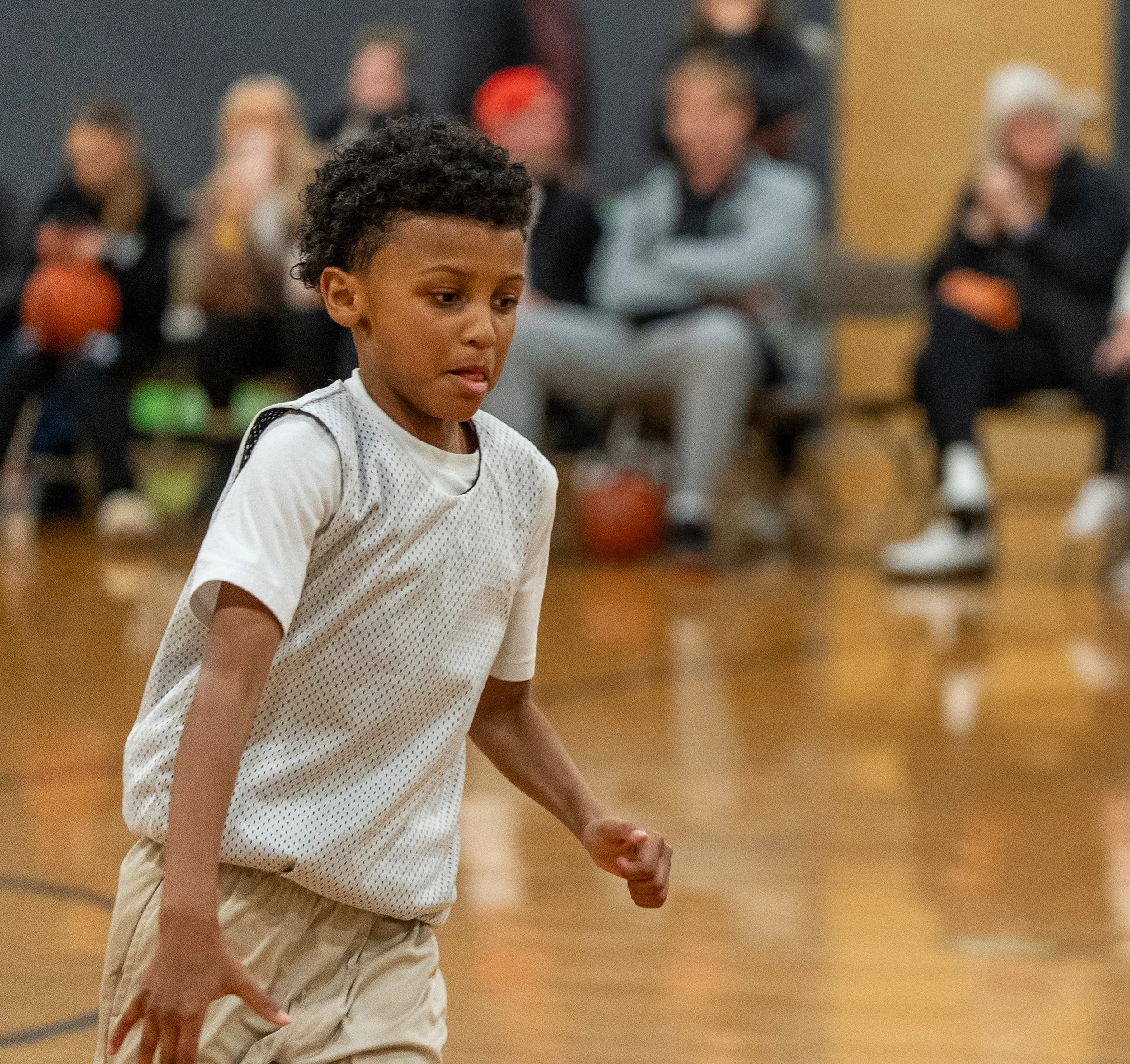 A young boy in a gymnasium, wearing a white sports jersey, appears to be focused or in thought during a basketball game or practice.
