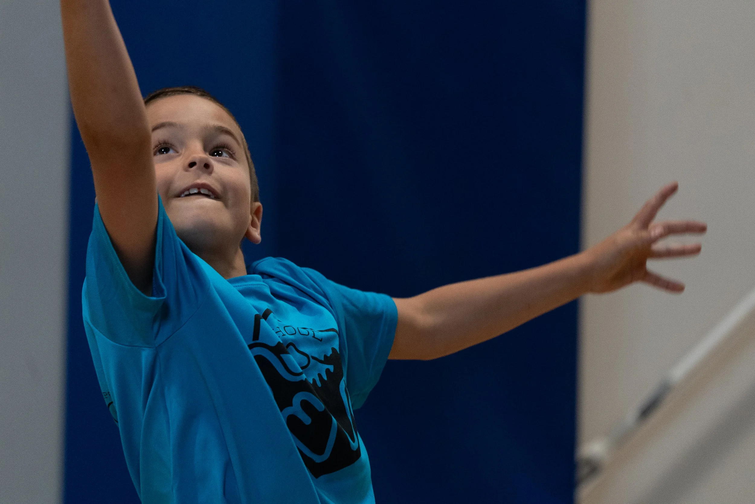 A young boy with short brown hair wearing a bright blue shirt reaches upward with both arms raised, looking up with an expression of excitement. The background features a dark blue wall on his left and a light-colored wall on his right.