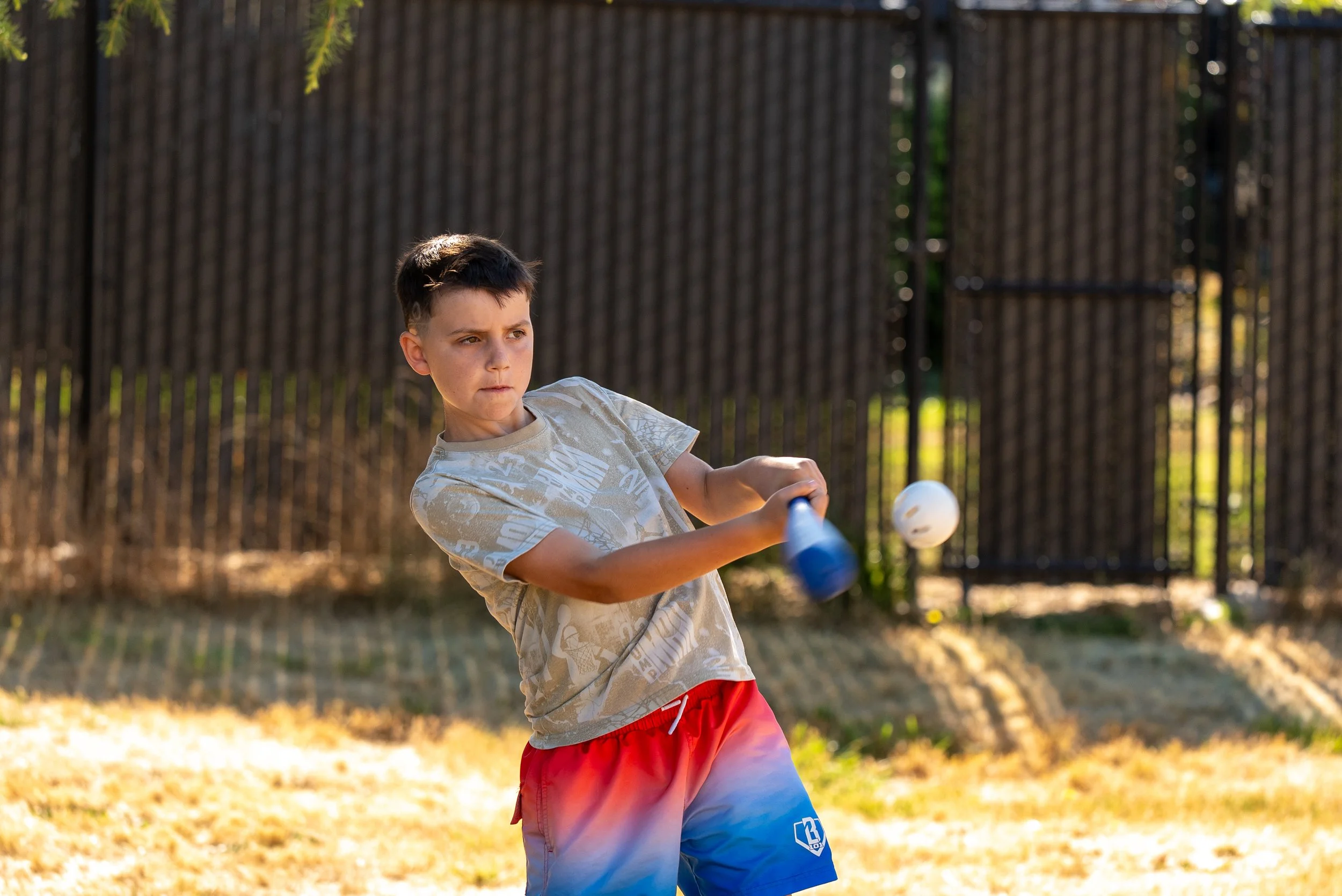 A young boy with short dark hair playing with a plastic bat and ball outdoors on a sunny day, with a wooden fence in the background.