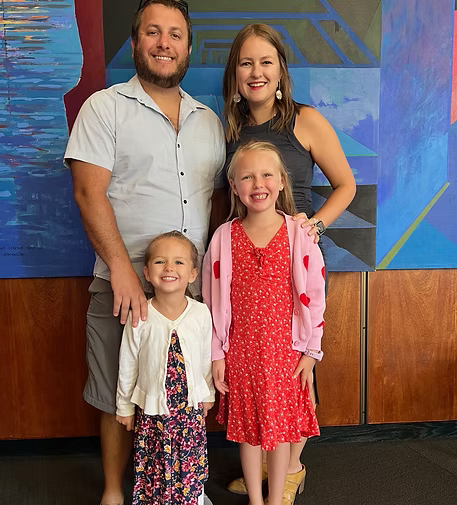 A family of four posing together indoors, with a colorful abstract mural in the background. The father has a beard and is wearing a white shirt. The mother has shoulder-length hair and is wearing a black sleeveless dress. The two daughters are in front, one wearing a white cardigan and a floral dress, the other in a red dress with a pink cardigan covered in red hearts.