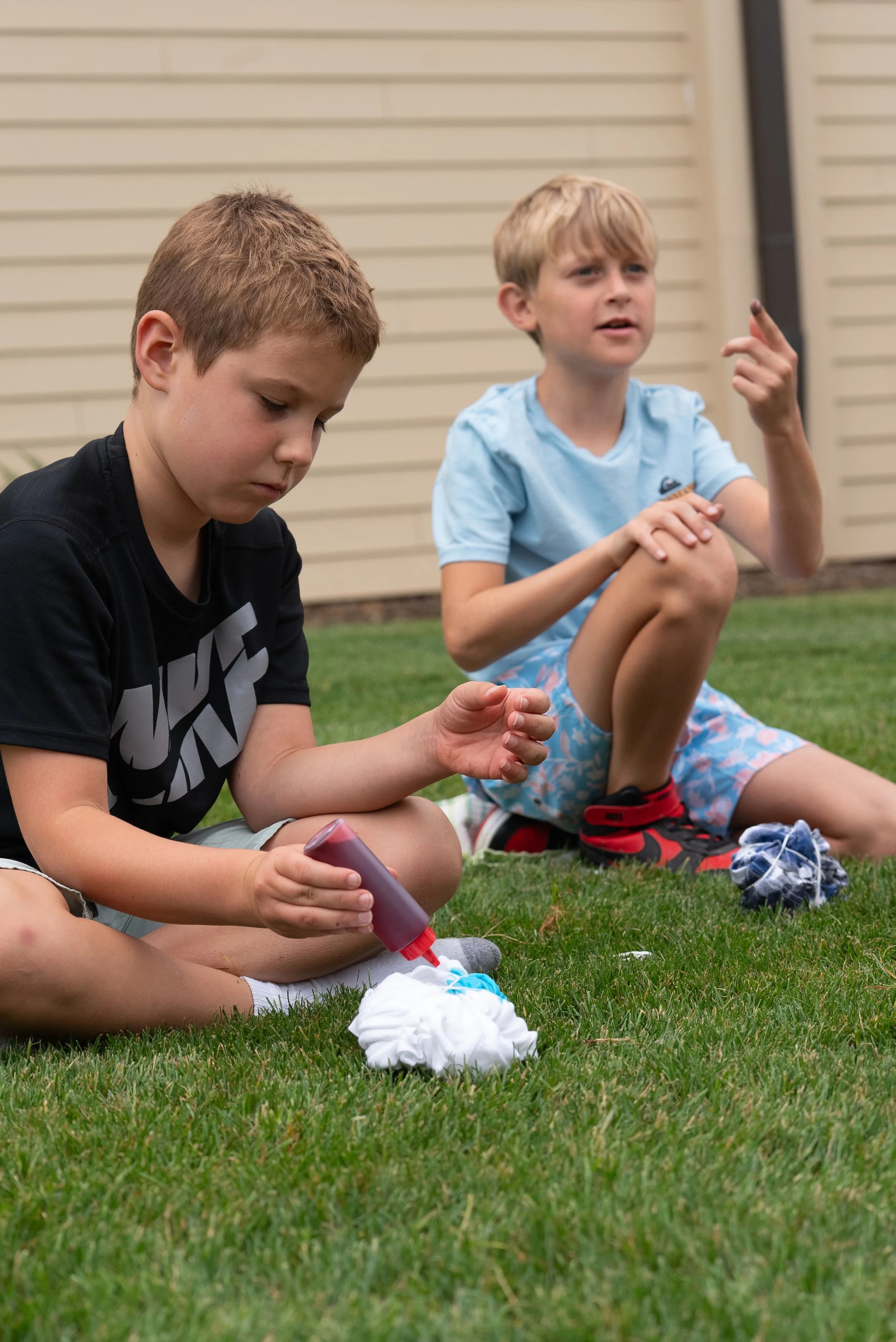 Two young boys sitting on the grass outside, one holding a bottle of white glue, and the other looking and gesturing with his hand, with clothes, shoes, and a pile of cloth or paper nearby.