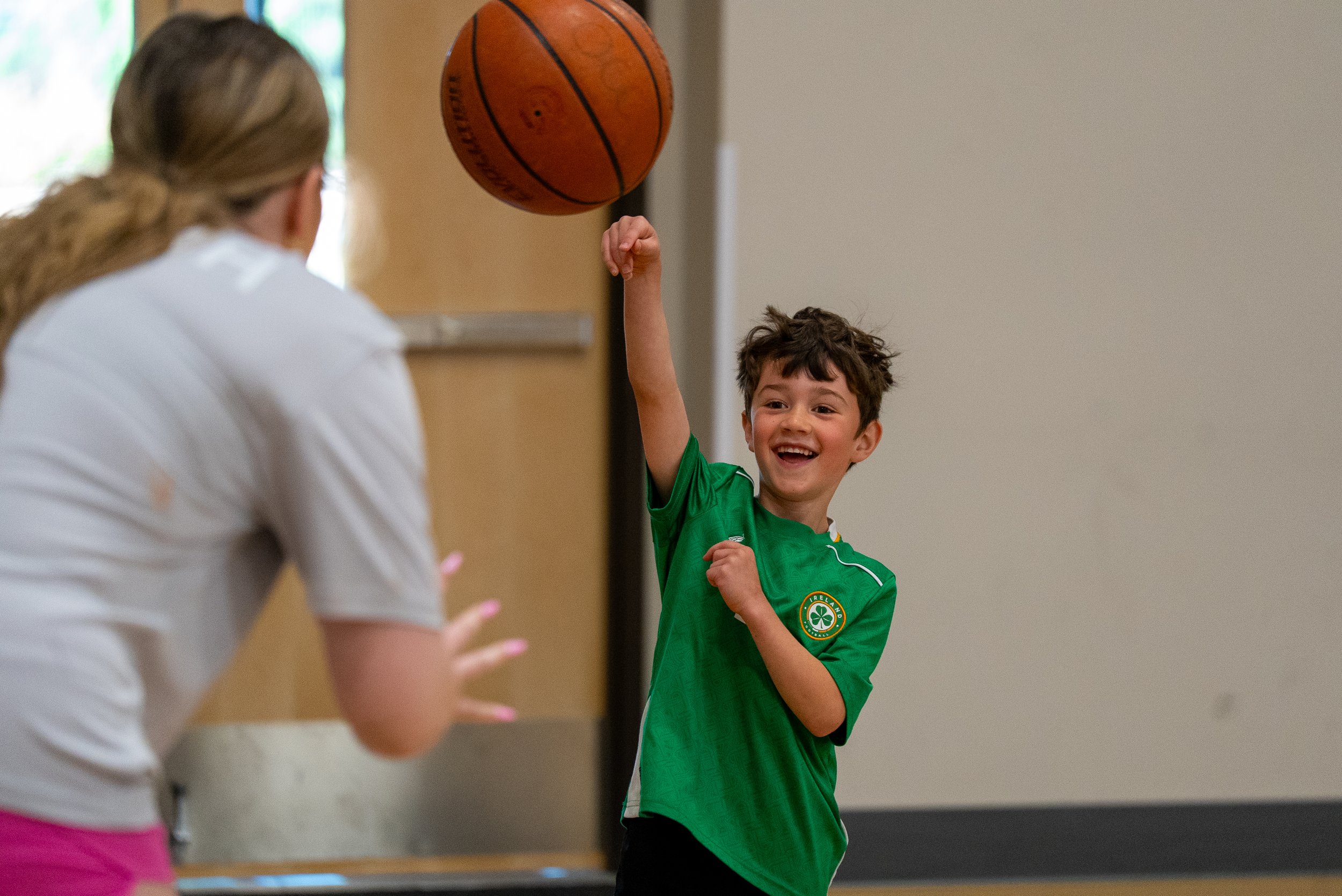 A young boy wearing a green sports jersey with a shamrock emblem is smiling and raising his arm to hit a basketball. An adult woman with light brown hair tied back, dressed in a white shirt, is facing him but is out of focus. The scene appears to be indoors, possibly in a gym or recreational area.