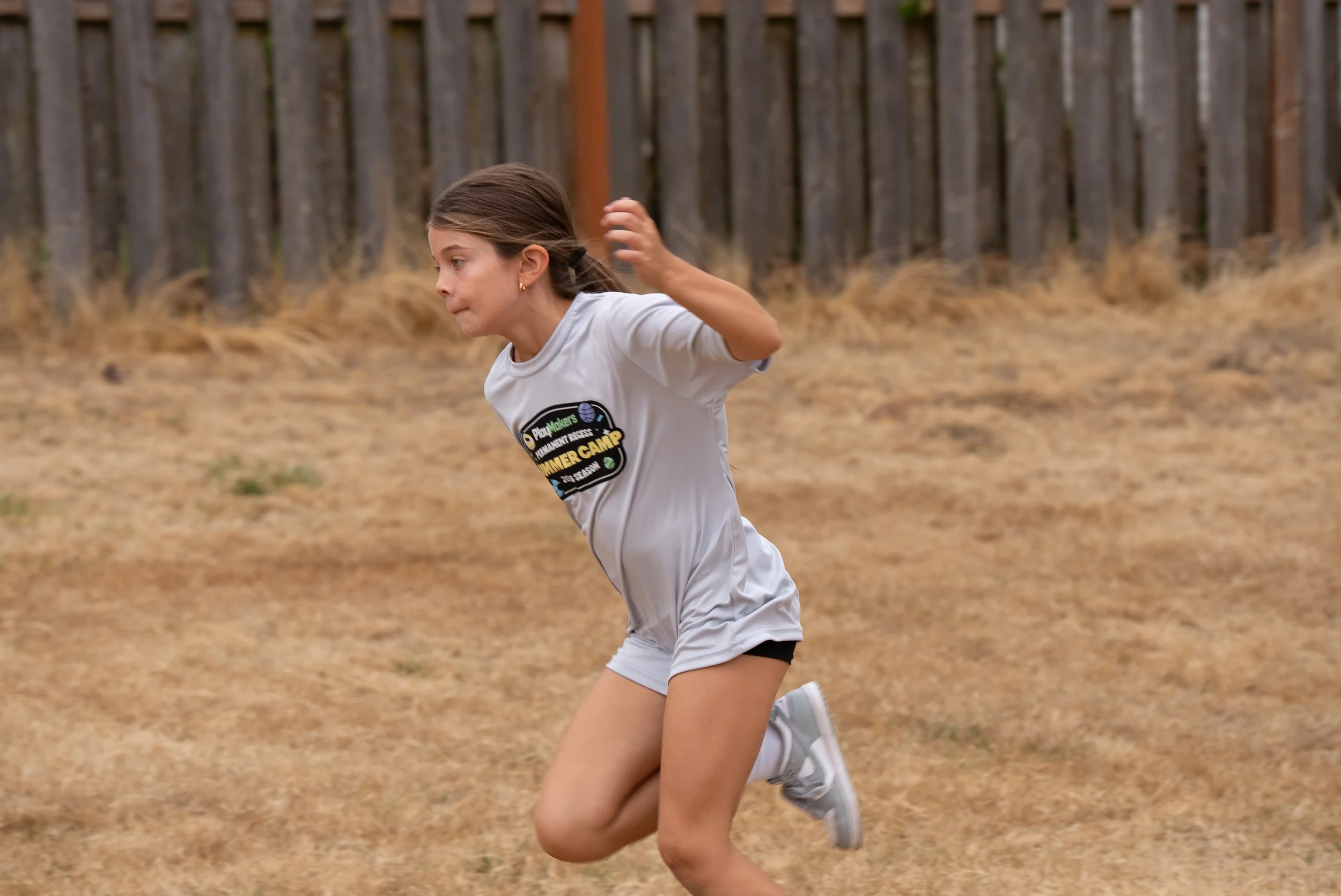 A young girl running outdoors on a dry, grassy field with a wooden fence in the background, wearing a white T-shirt and shorts.