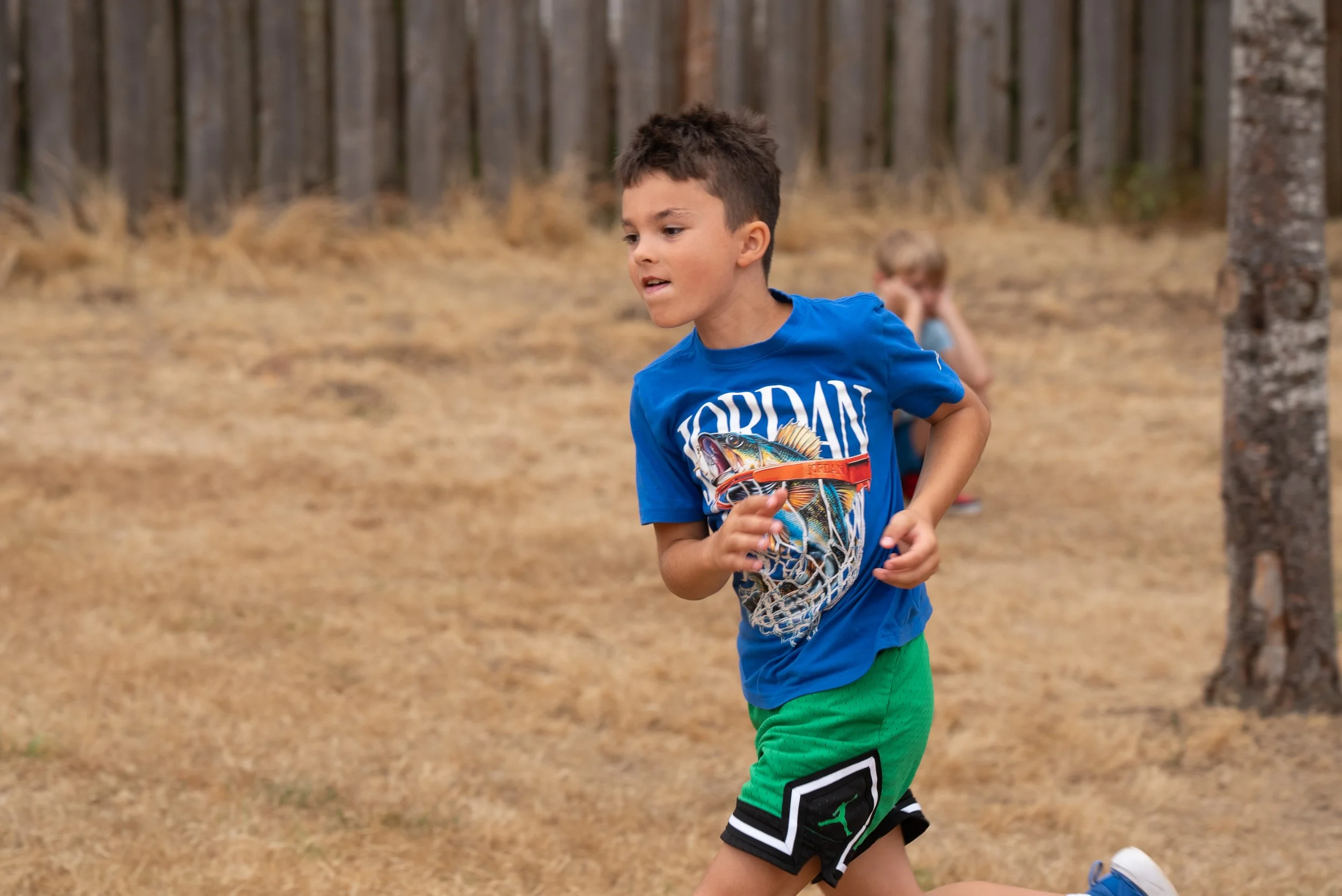 A young boy with short dark hair running outdoors, wearing a blue t-shirt with a colorful fish graphic and the word 'JORDAN,' and green shorts with black and white trim, in a wooded area with dry grass and trees in the background.
