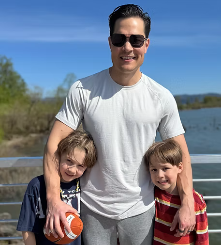 A smiling man wearing sunglasses standing outdoors on a bridge with two young boys, all enjoying a sunny day by a river or lake with trees in the background.