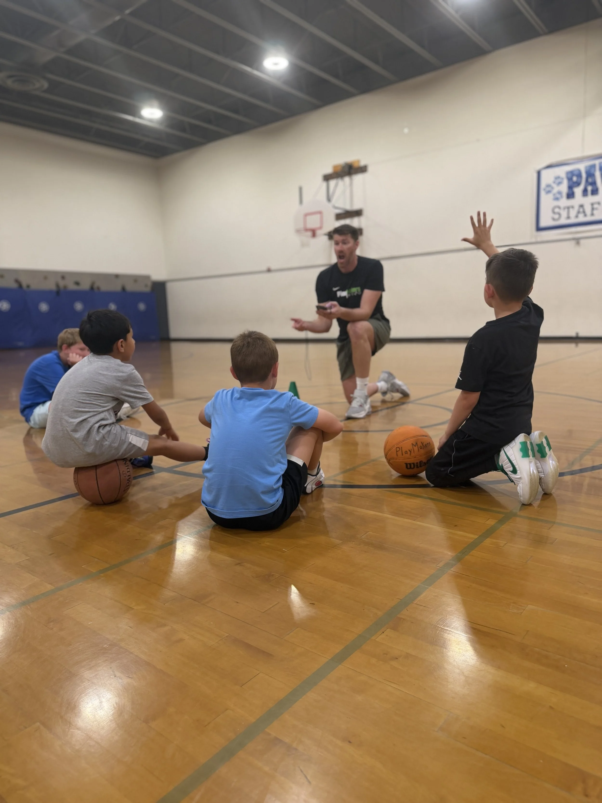 A group of young boys sitting on the gym floor during a basketball camp, listening to an instructor who is explaining something, with one boy raising his hand.