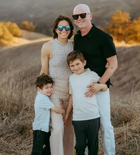 A smiling family of four standing outdoors in a grassy field with rolling hills and trees with autumn foliage in the background.