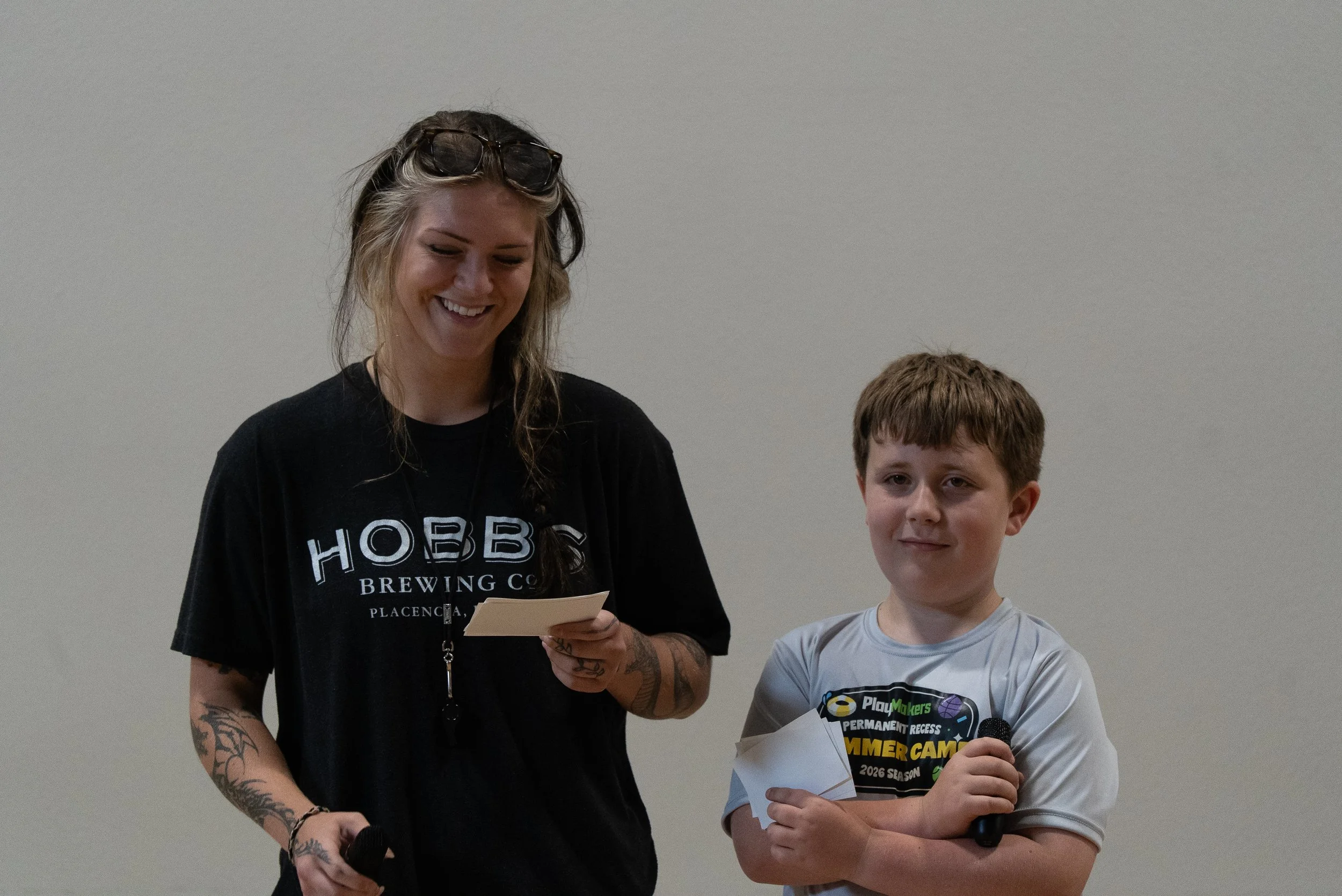 A woman with glasses on her head, tattoos, and a black t-shirt, smiling and holding a piece of paper, standing next to a young boy with brown hair, wearing a white t-shirt, holding a microphone, standing against a plain light-colored wall.