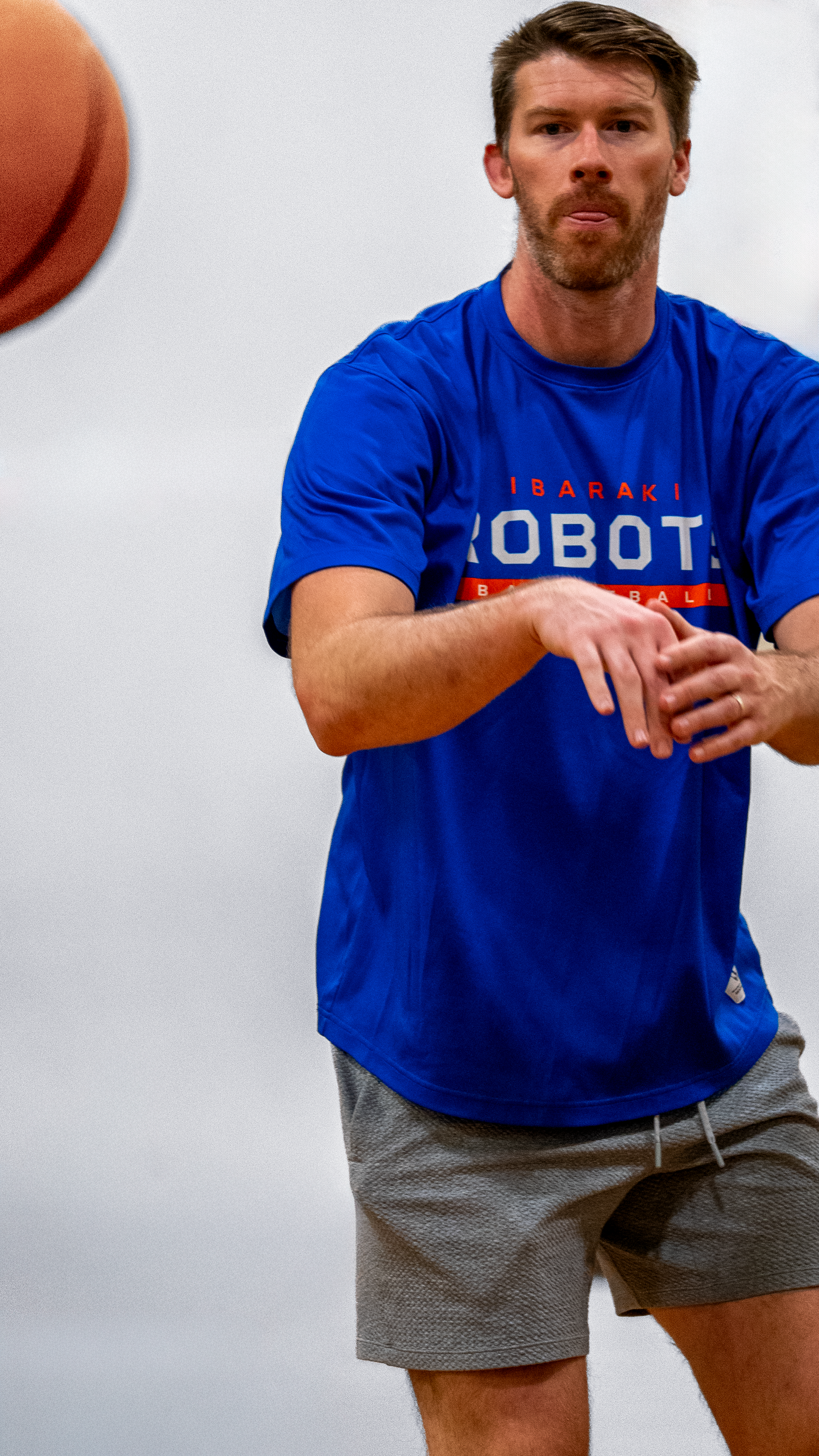 A man in a blue T-shirt with the words 'IBARAKI ROBOT' and gray shorts playing basketball, with a basketball visible near his head.