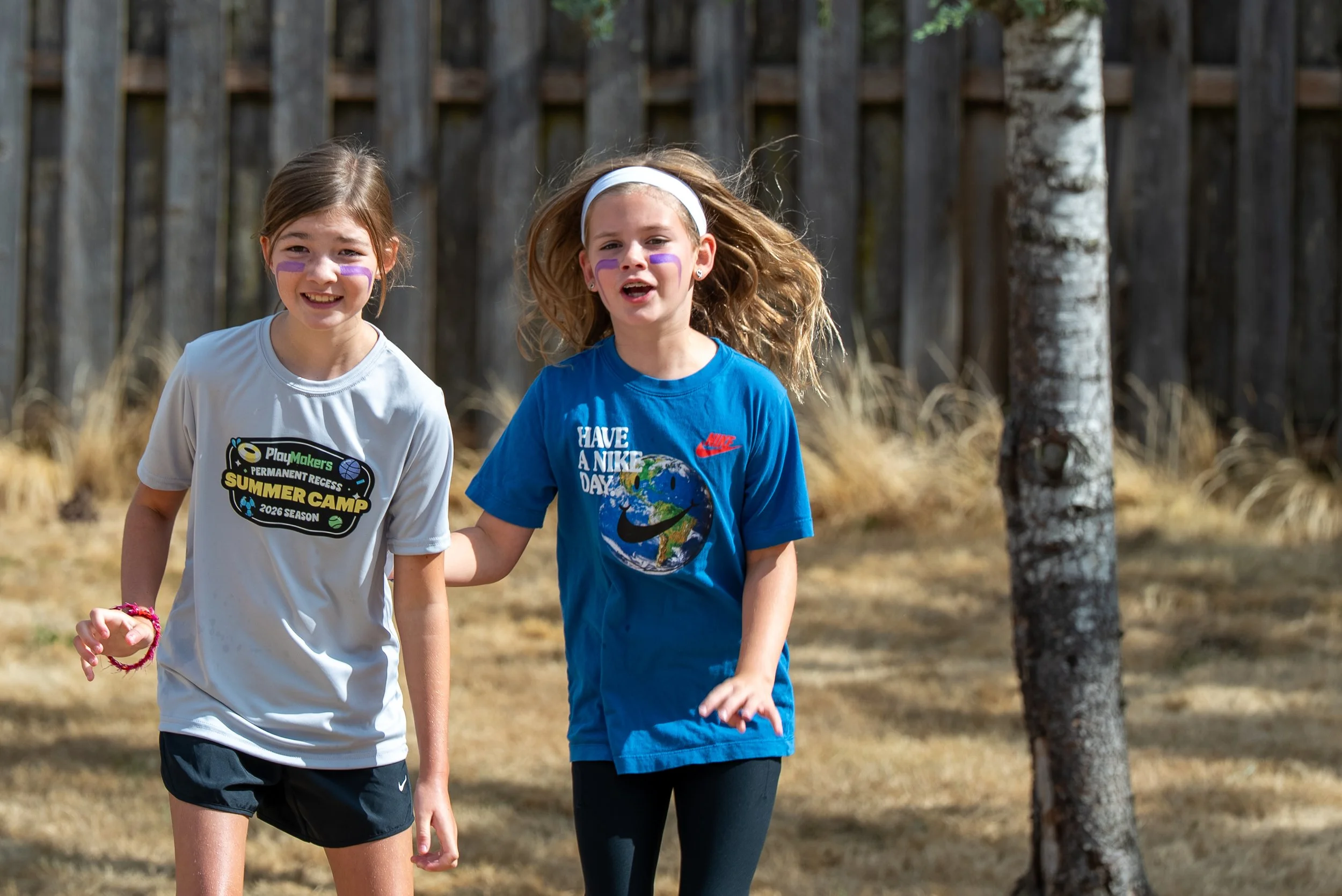 Two young girls with face paint, running outdoors in a wooded area during daytime. One girl wears a white T-shirt with a logo and black shorts, and the other wears a blue T-shirt with a globe print and black leggings.