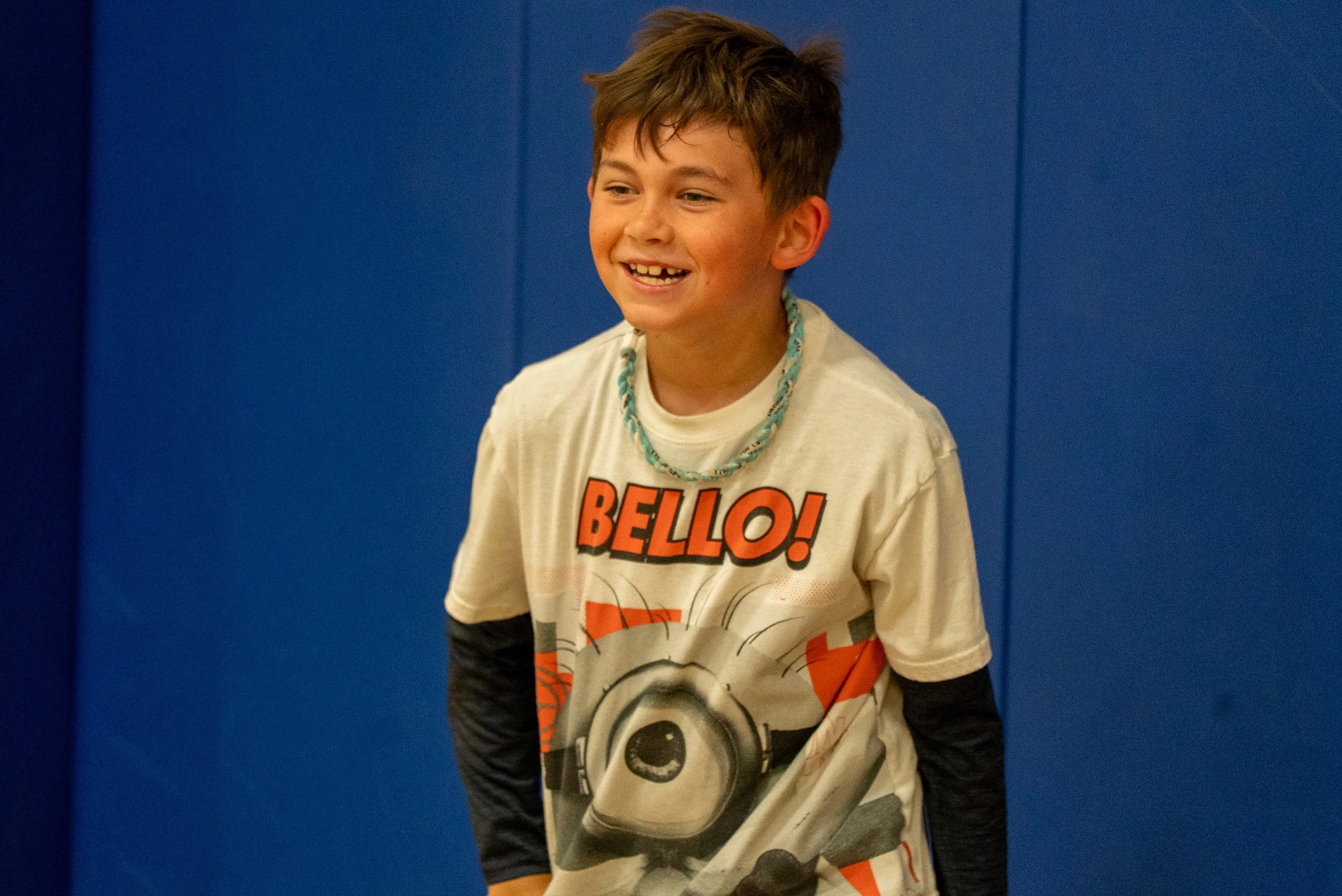 A young boy with short brown hair smiling, wearing a graphic t-shirt featuring a character and the word 'Bello!', a chain necklace, and long-sleeve black shirt underneath. He is standing against a blue wall.