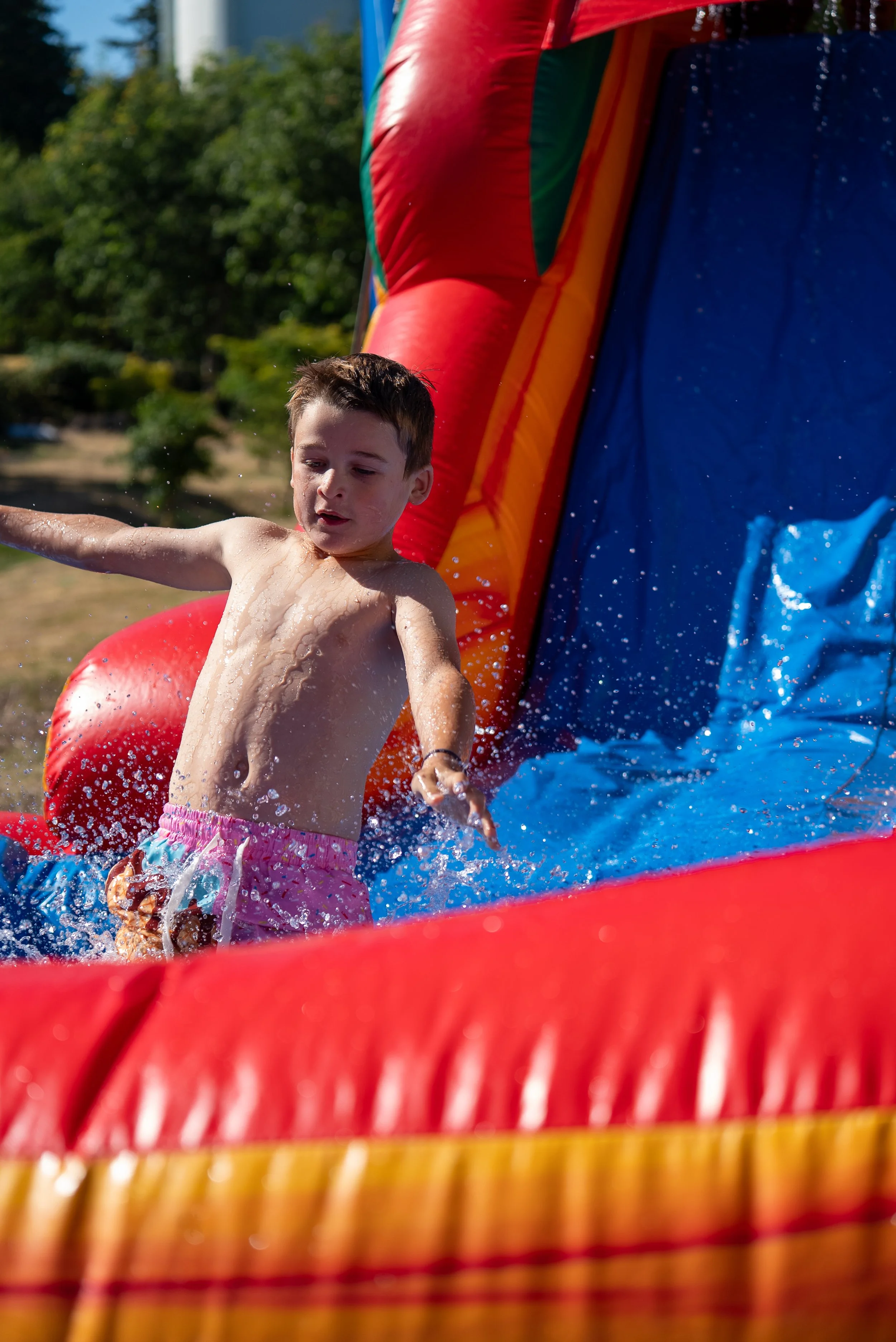 A young boy wearing pink shorts is sliding down a colorful inflatable water slide outside on a sunny day, with trees and a partly cloudy sky in the background.