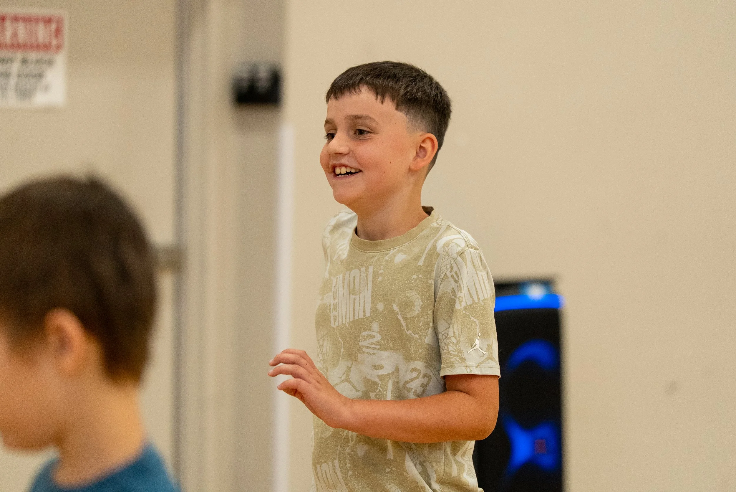 A young boy with short dark hair, smiling, wearing a beige T-shirt with graphic text, standing indoors, possibly in a classroom or community center.